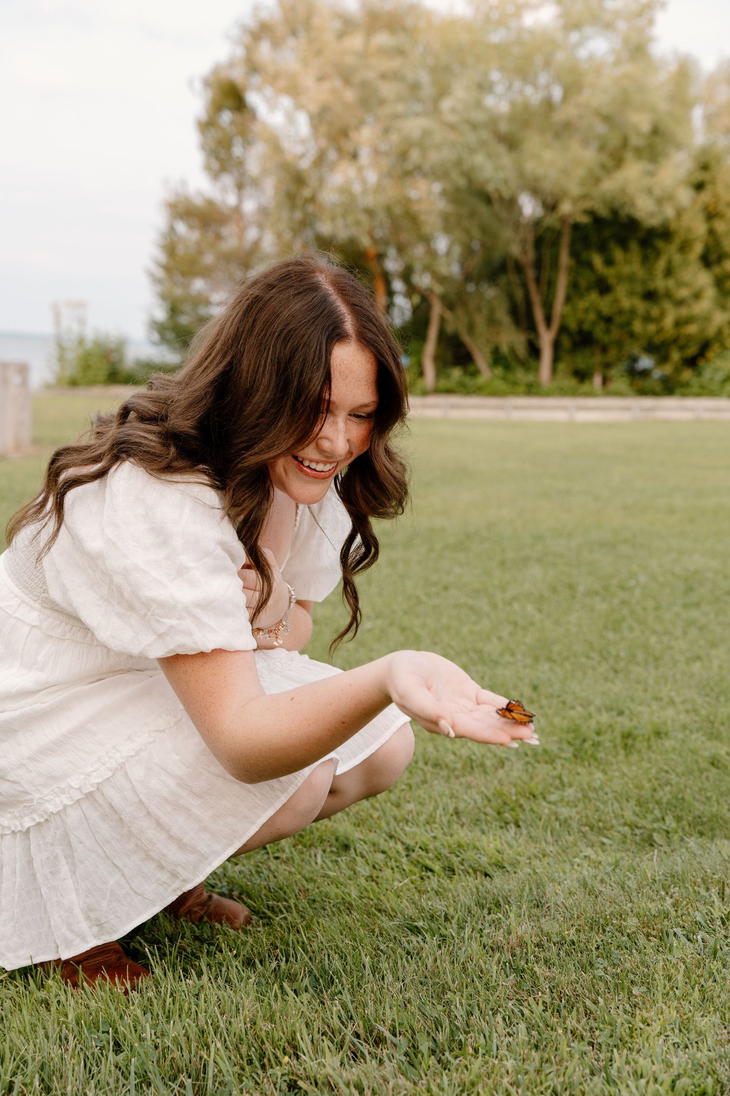 A smiling brunette woman in a white dress crouches on green grass, holding a small butterfly in her hand.