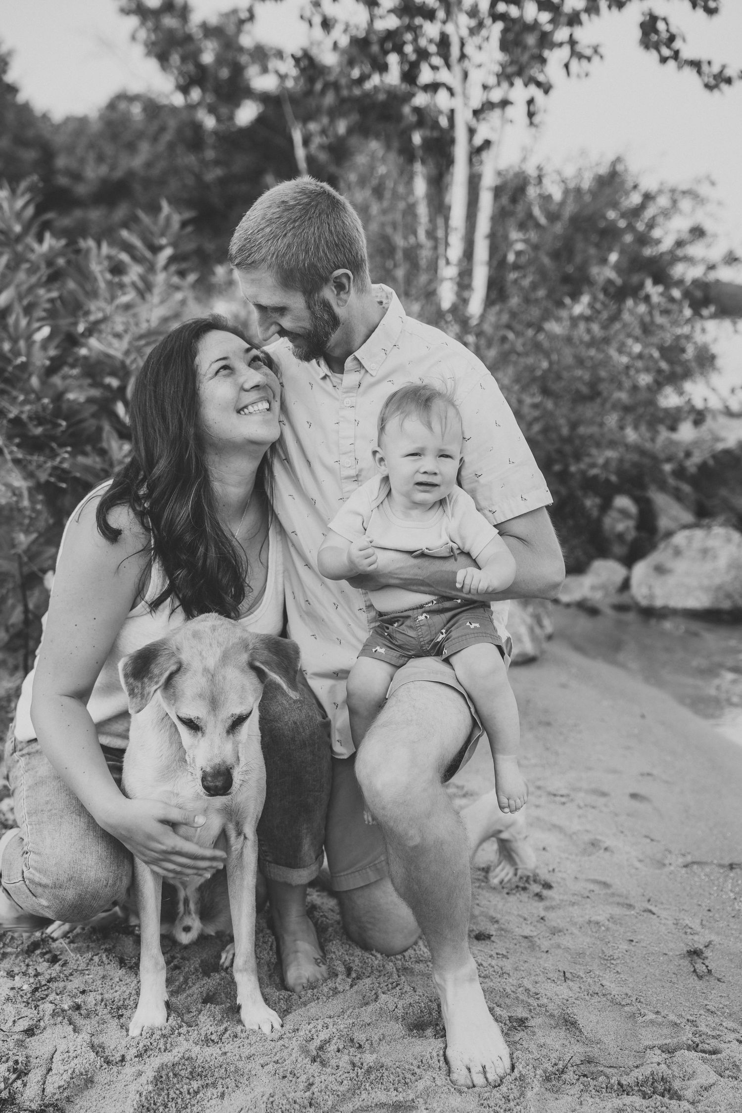 Black and white family photo of couple with baby and dog on sandy beach surrounded by trees.