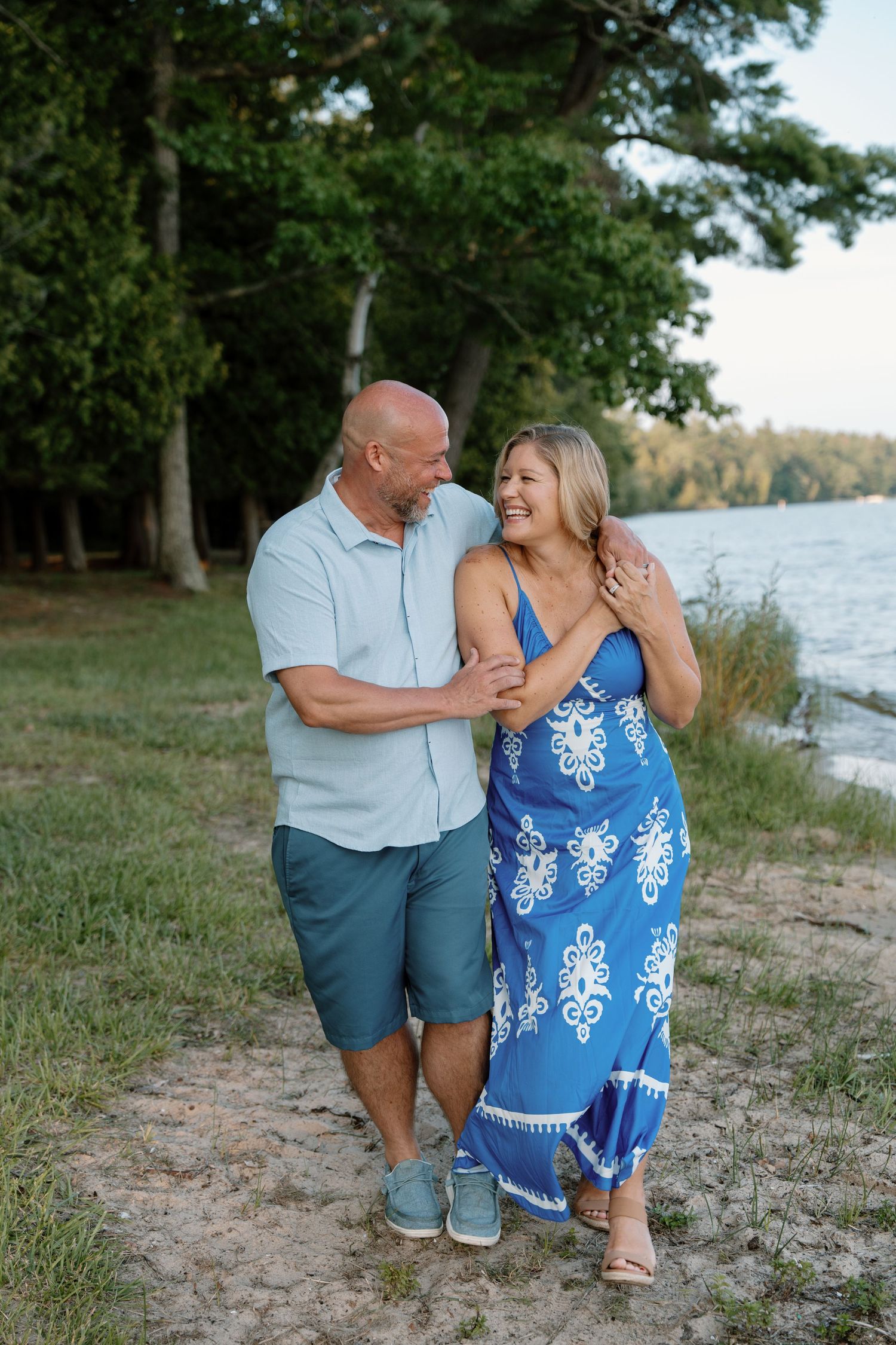 A smiling couple embraces on a sandy lakeside path surrounded by lush green trees at golden hour.