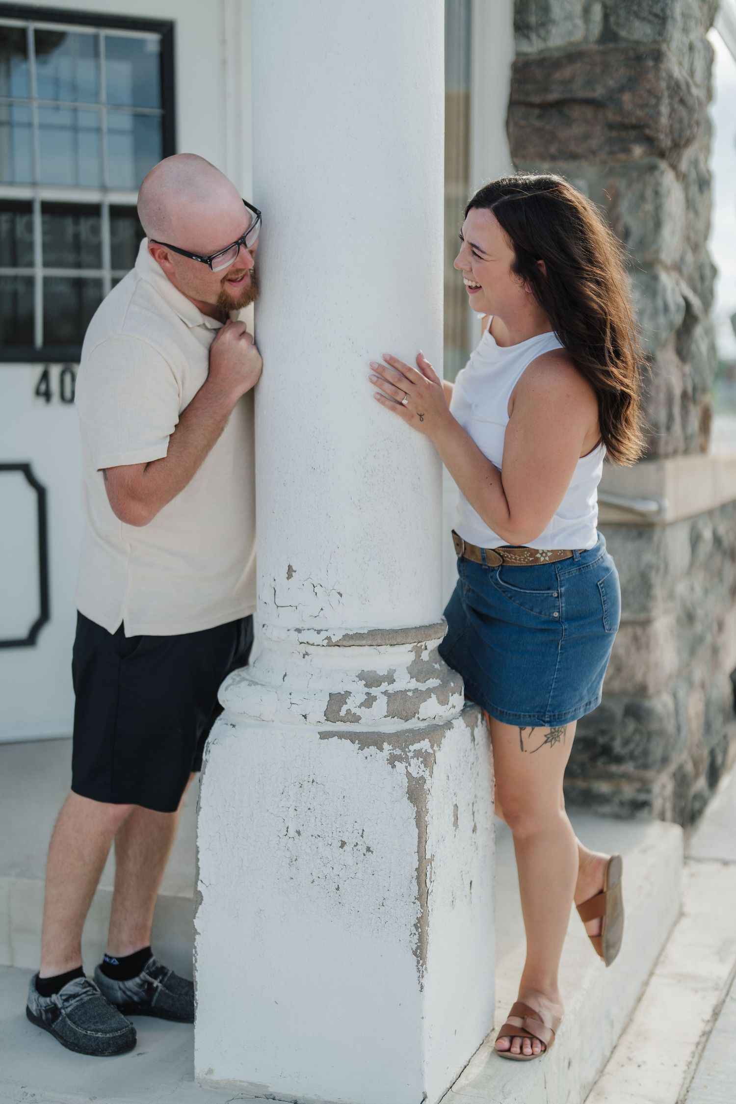 A bald man and brunette woman laugh together beside a white column on a building's porch.