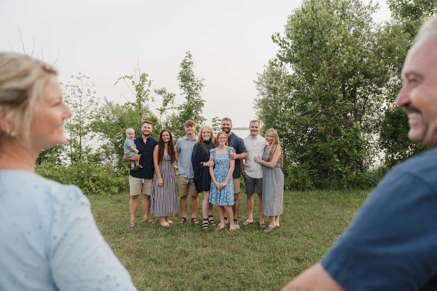 Large family group photo outdoors on green grass with trees, posing together for a portrait session.