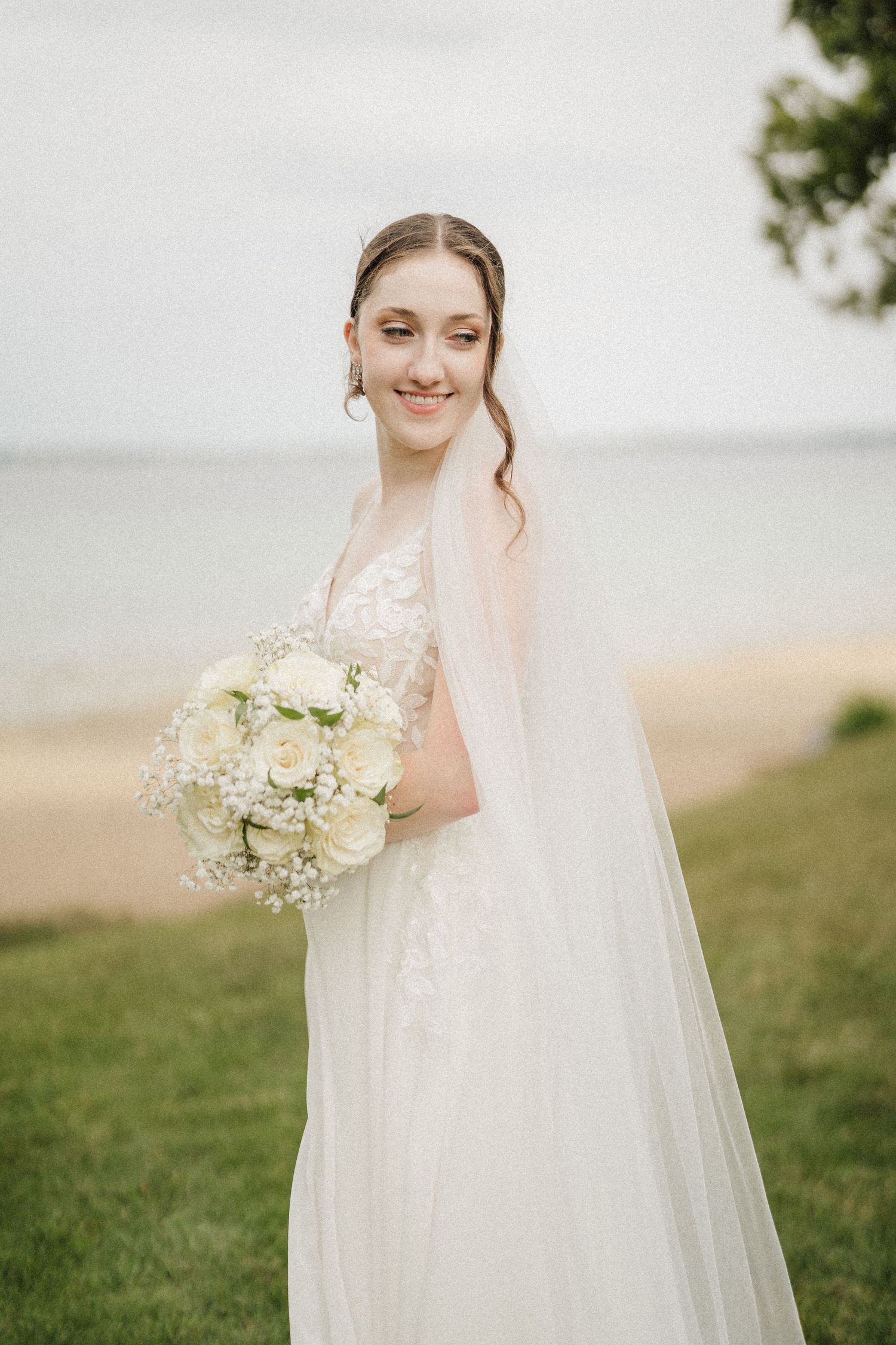 A bride in a flowing white wedding dress holds a delicate bouquet of white flowers against a soft outdoor background.