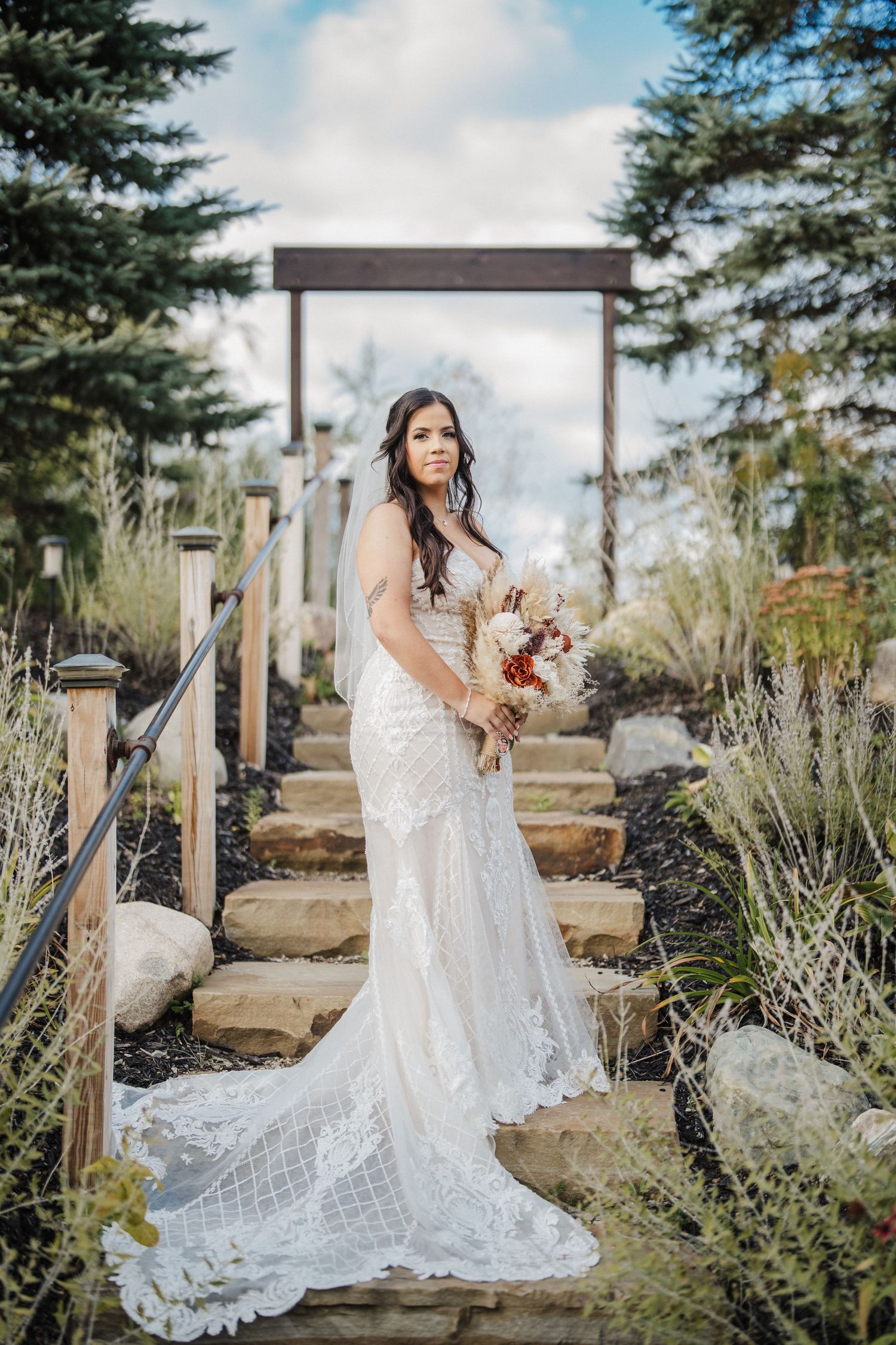 Bride in lace wedding gown poses among ornamental grasses and garden landscaping.