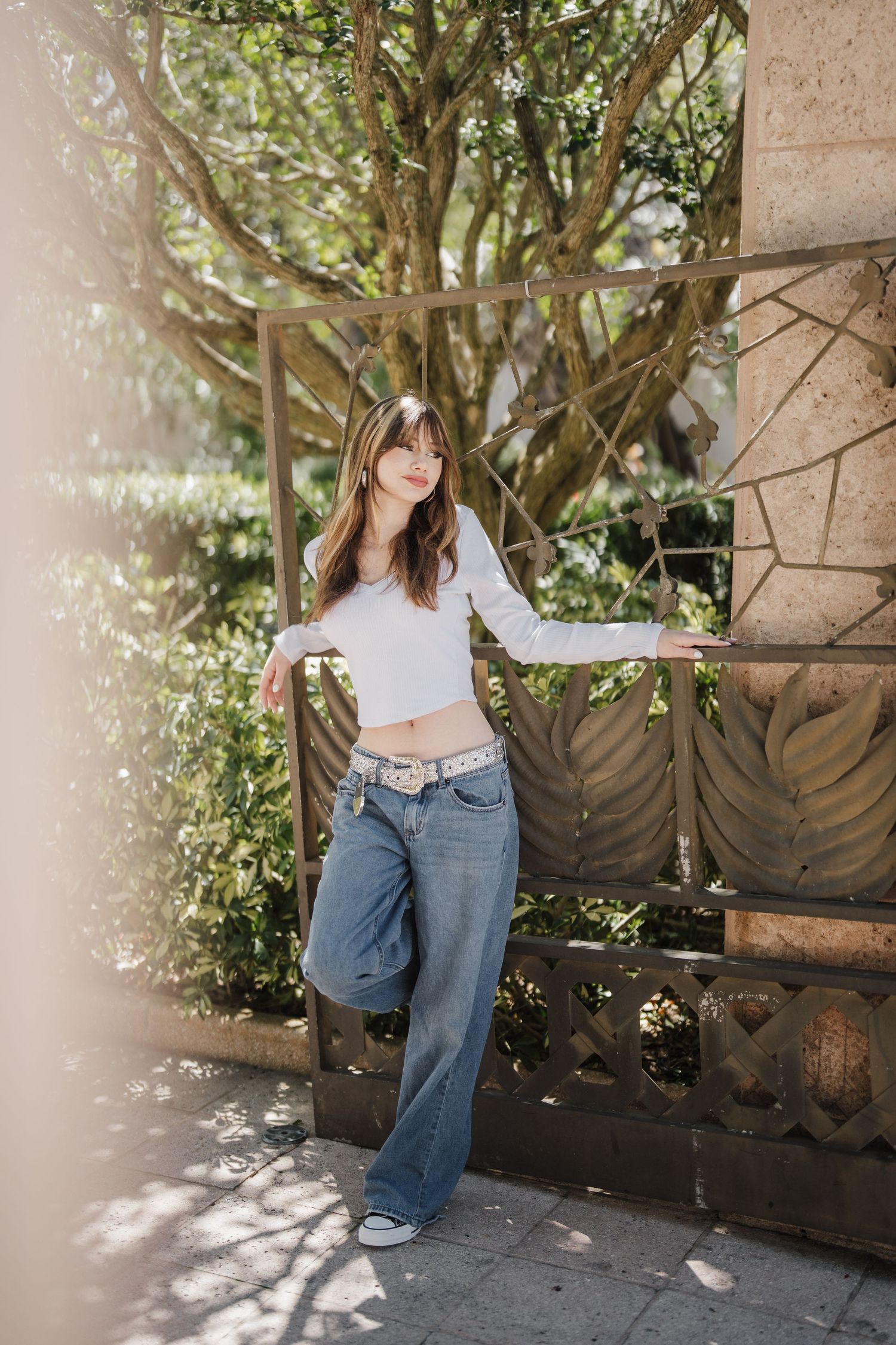 A casual outdoor fashion pose featuring wide-leg jeans and a white crop top against a garden backdrop.