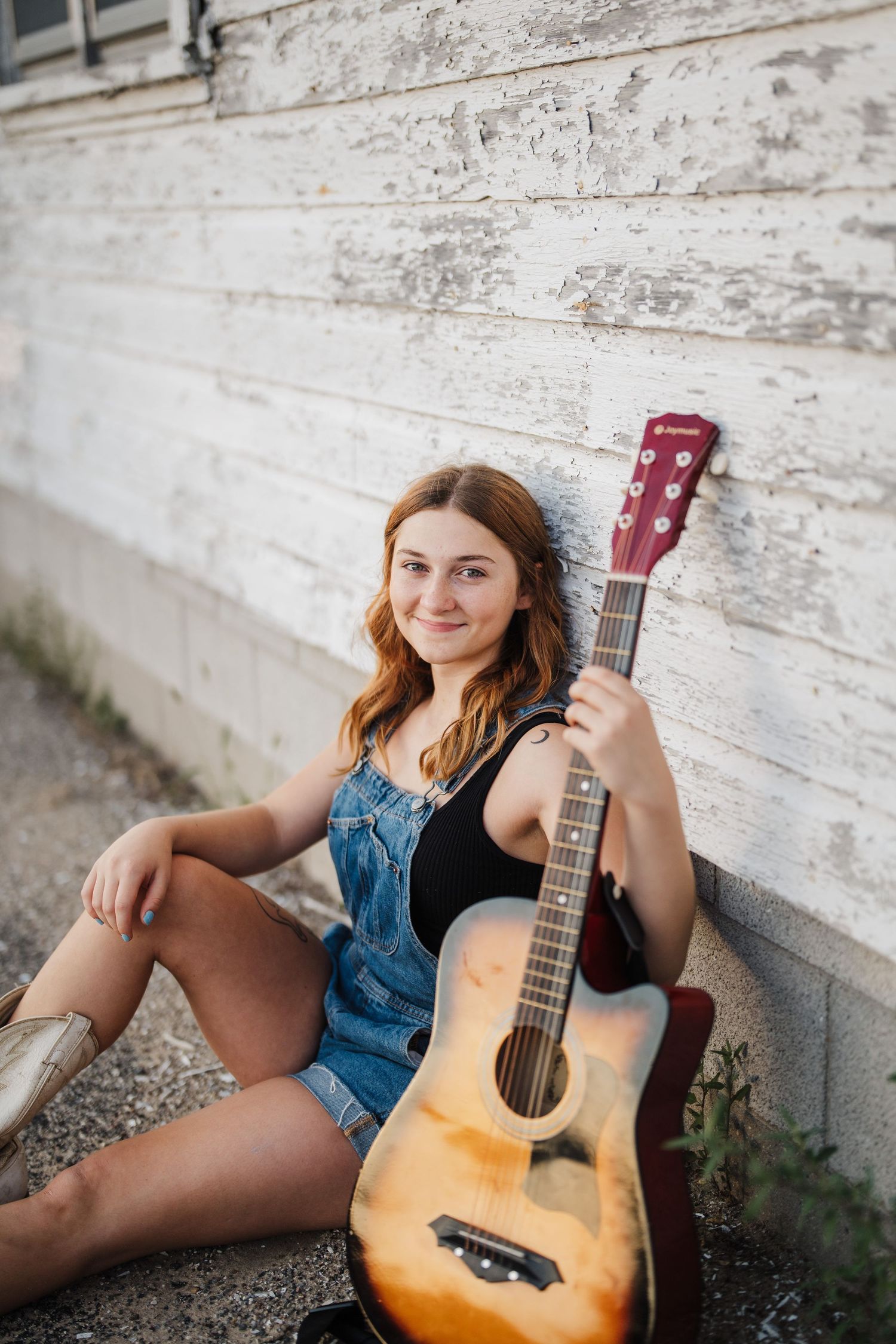 A person in denim sits against a rustic white wall while playing an acoustic guitar in a casual outdoor setting.