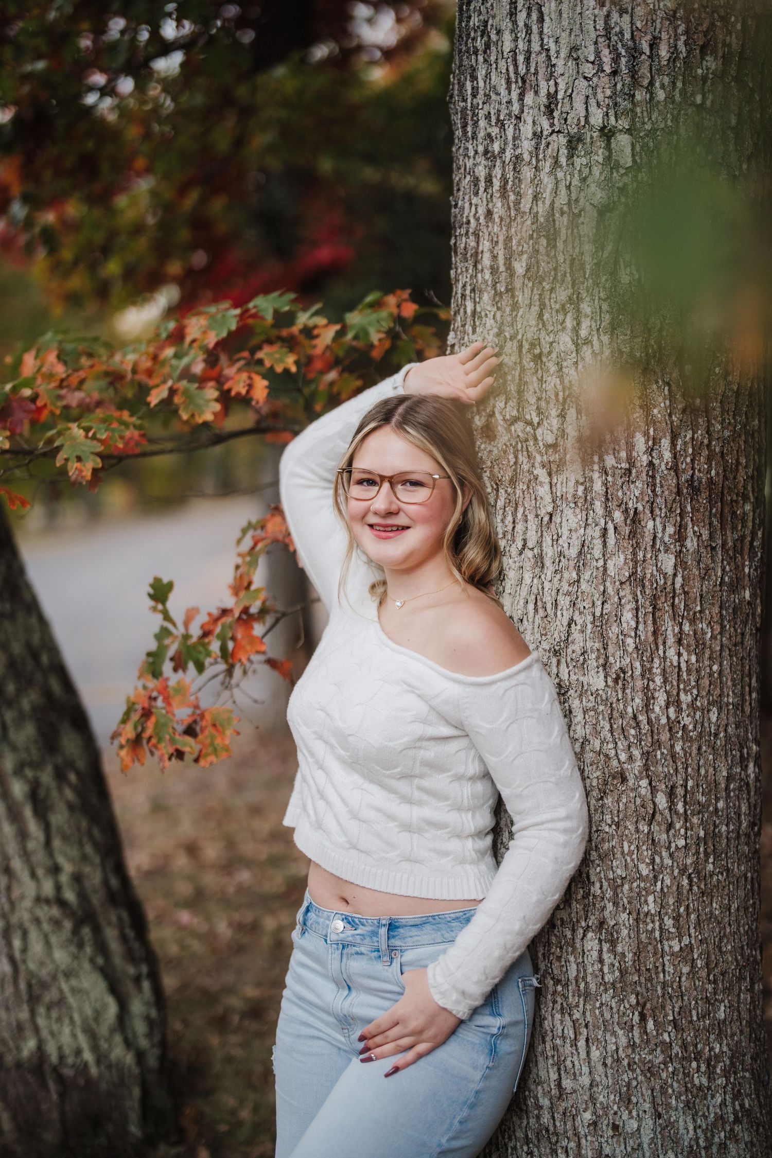 Outdoor autumn portrait session featuring someone in a white sweater and jeans posing against a tree trunk with fall foliage.