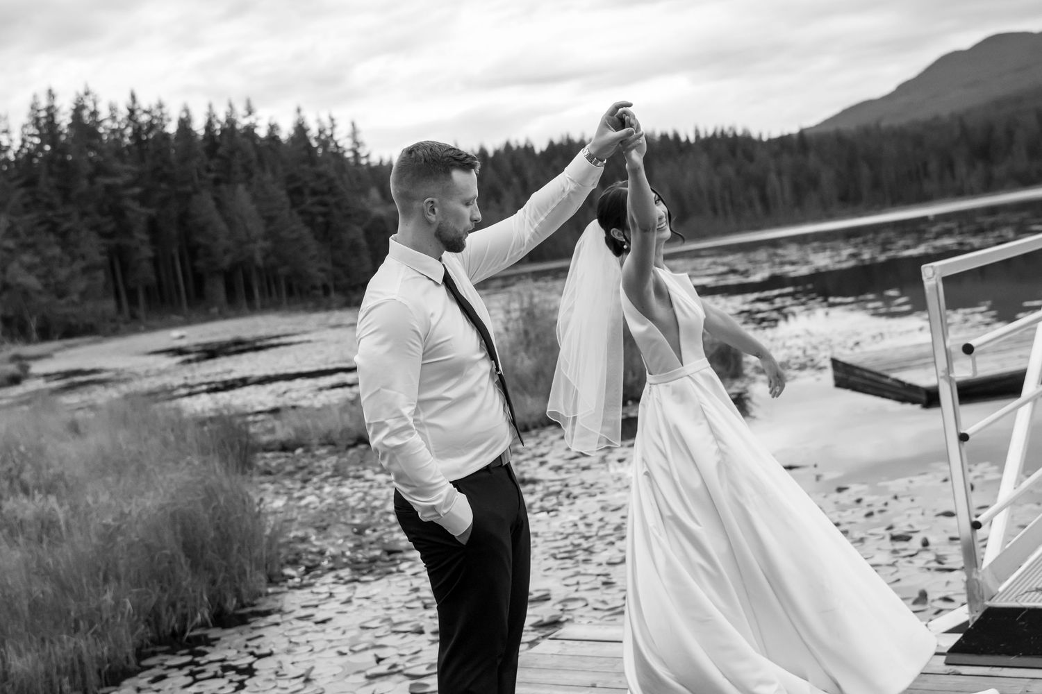 Black and white photo of a groom twirling his bride on a dock by a lake surrounded by pine trees.