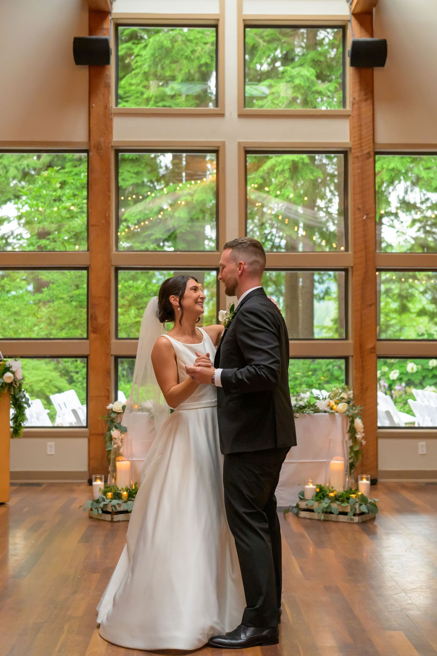 Bride and groom share first dance in elegant wood-paneled chapel with floor-to-ceiling windows and lush greenery backdrop.
