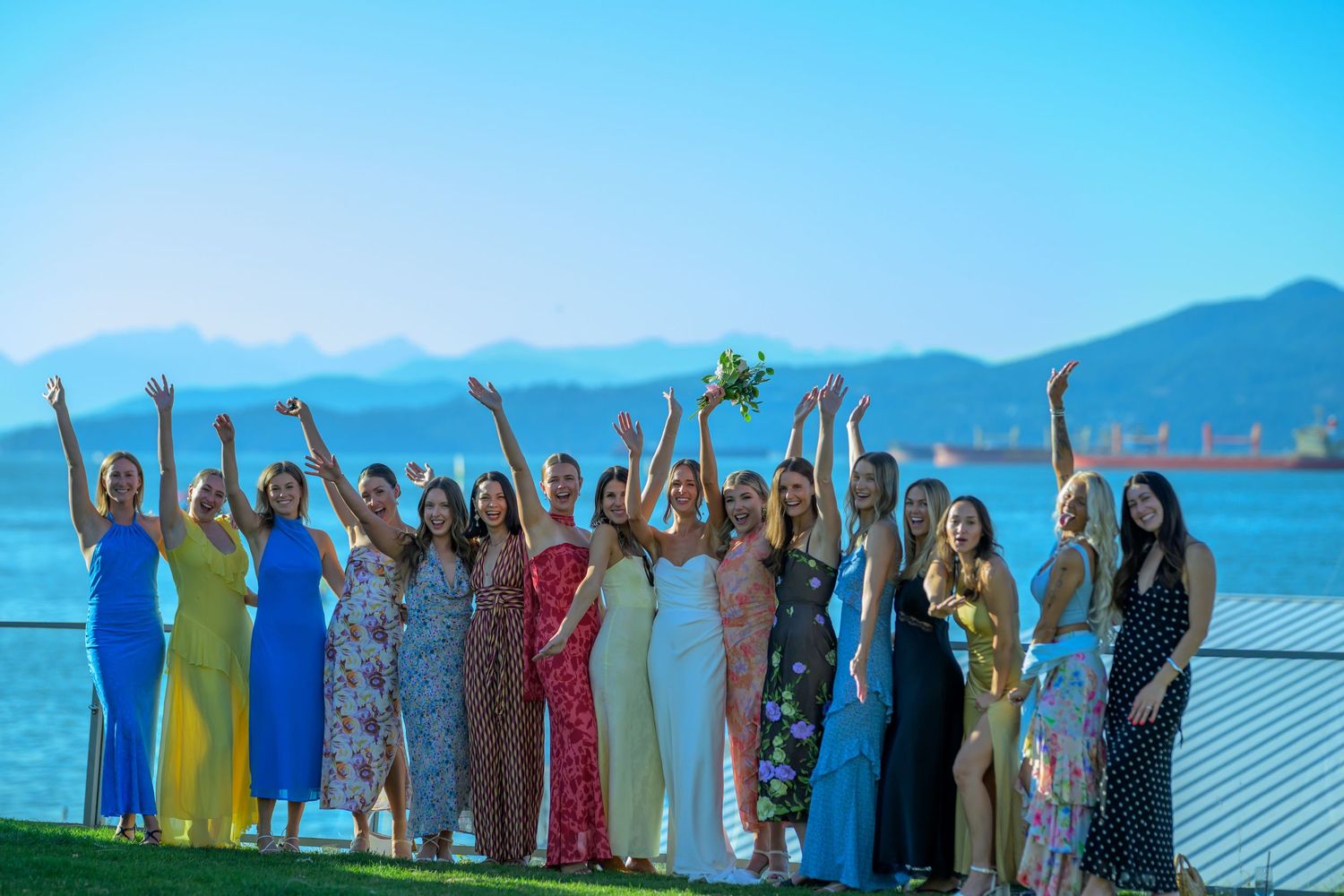 Wedding party group photo with colorful attire against mountain and ocean backdrop.