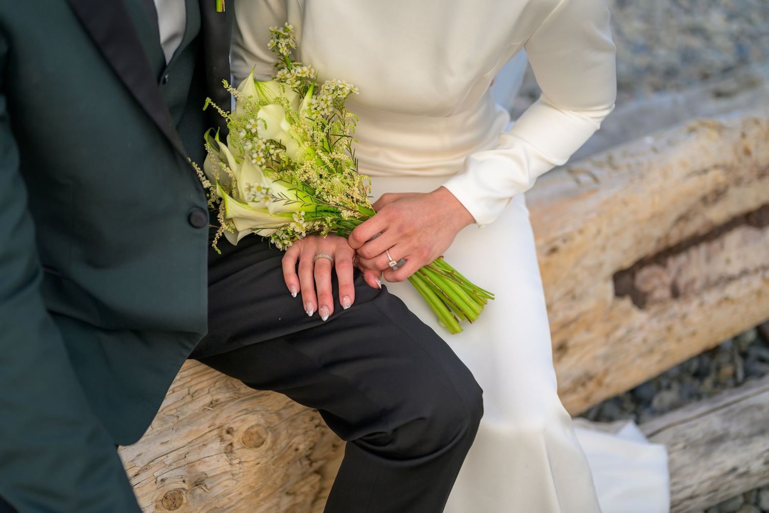 Close-up of wedding couple holding green and white bouquet while sitting on driftwood.