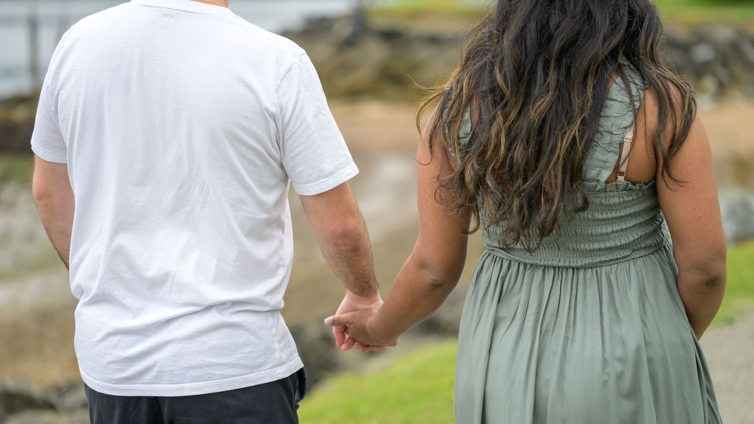 Couple holding hands walking outdoors near the ocean, man in white shirt and woman in green dress.