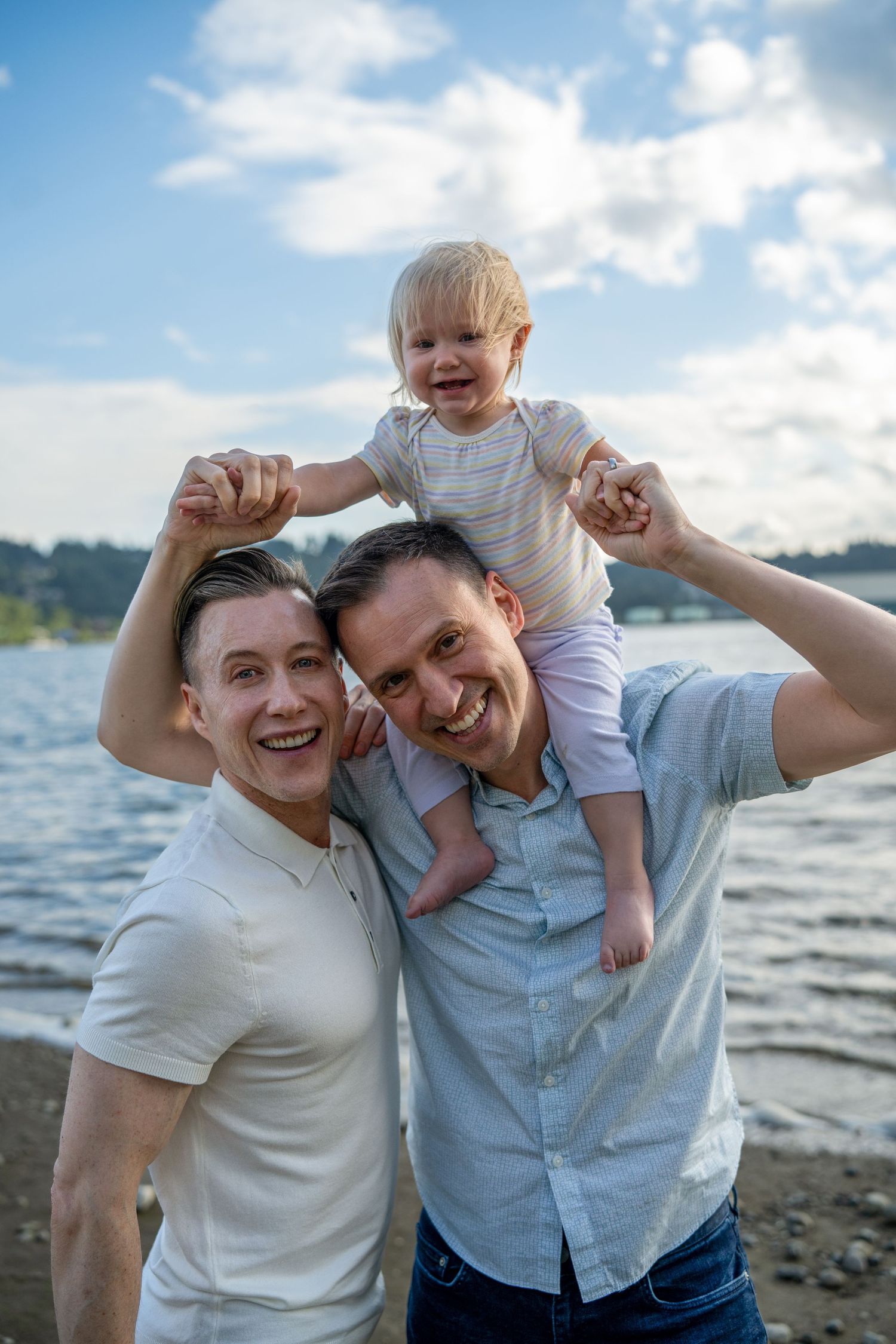 Two happy men smile at the camera on a beach, with a laughing toddler sitting on one man's shoulders.
