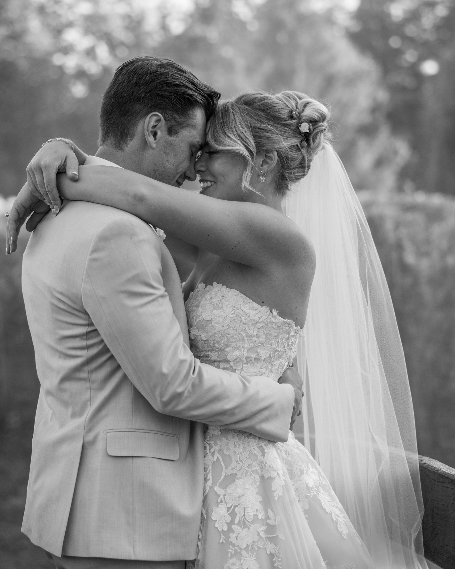 Black and white wedding photo of bride and groom embracing forehead to forehead, bride in lace gown with veil.