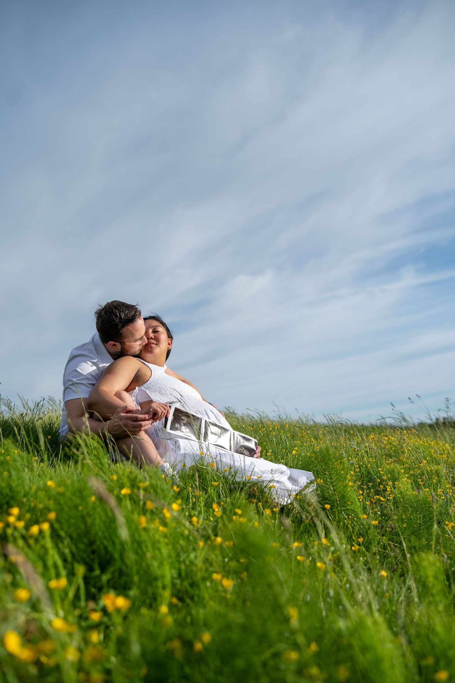 Couple sitting in a green meadow with yellow flowers, sharing a kiss while holding ultrasound photos on a sunny day.
