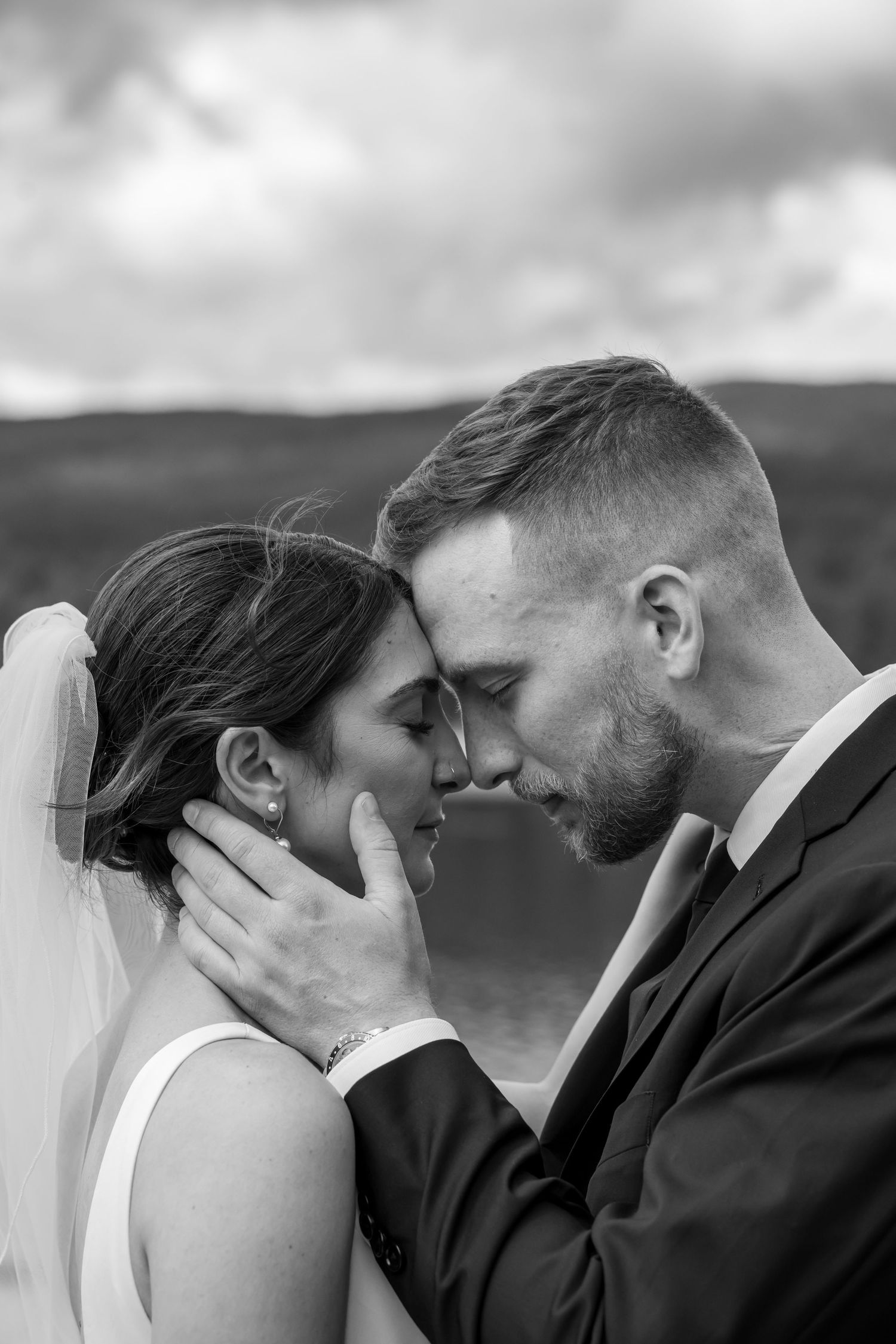 Black and white wedding photo of bride and groom touching foreheads tenderly outdoors with scenic hills backdrop.