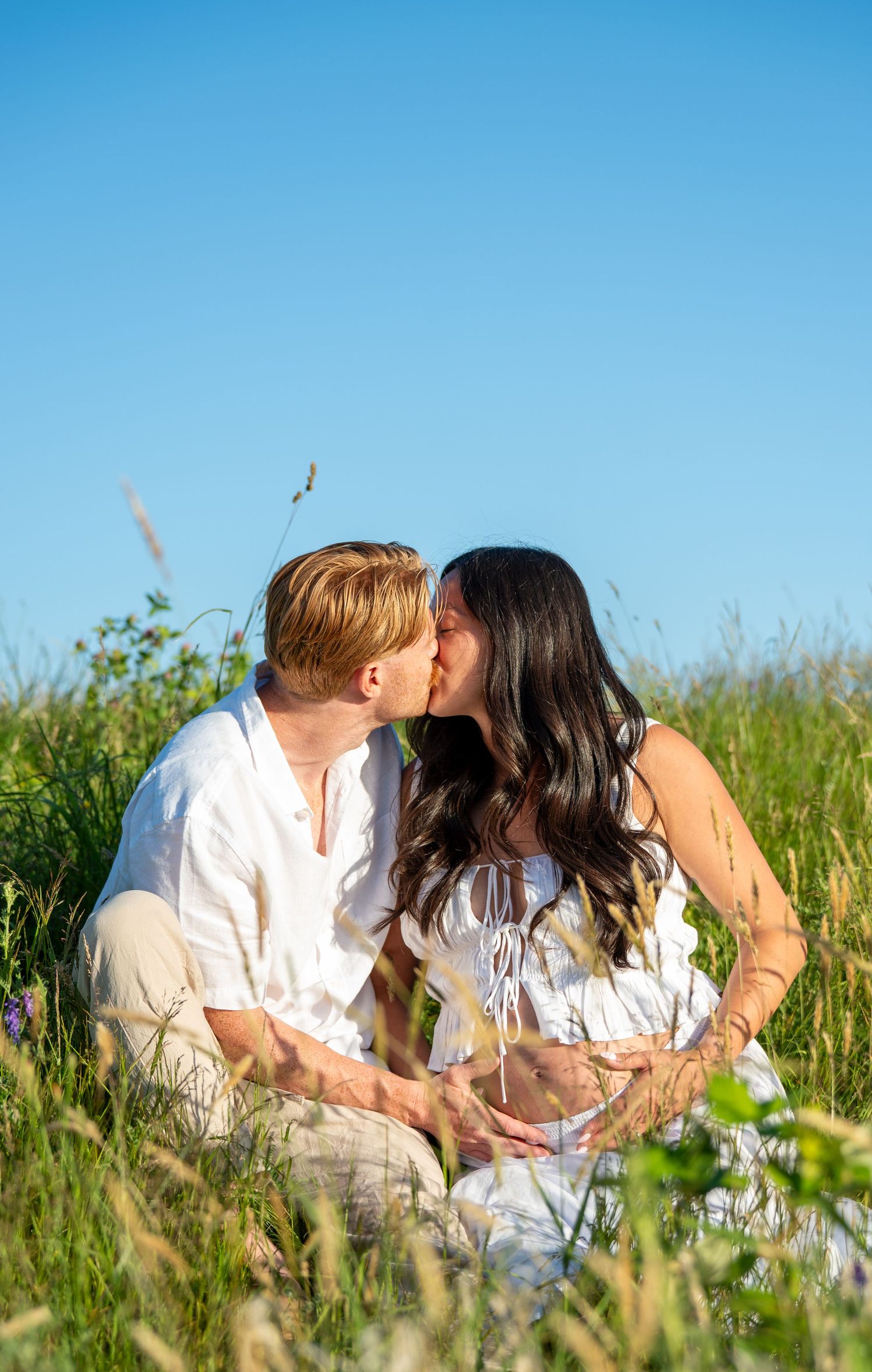 Couple kissing while sitting in a green meadow under a clear blue sky, both wearing white outfits.