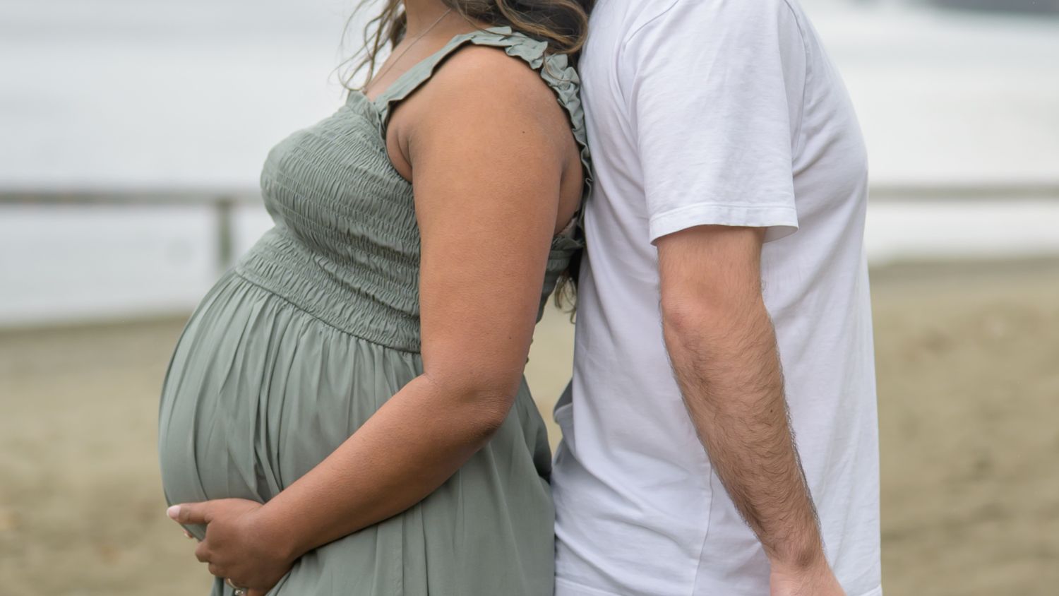 Pregnant woman in sage green dress with partner on beach, maternity photo shoot outdoors.