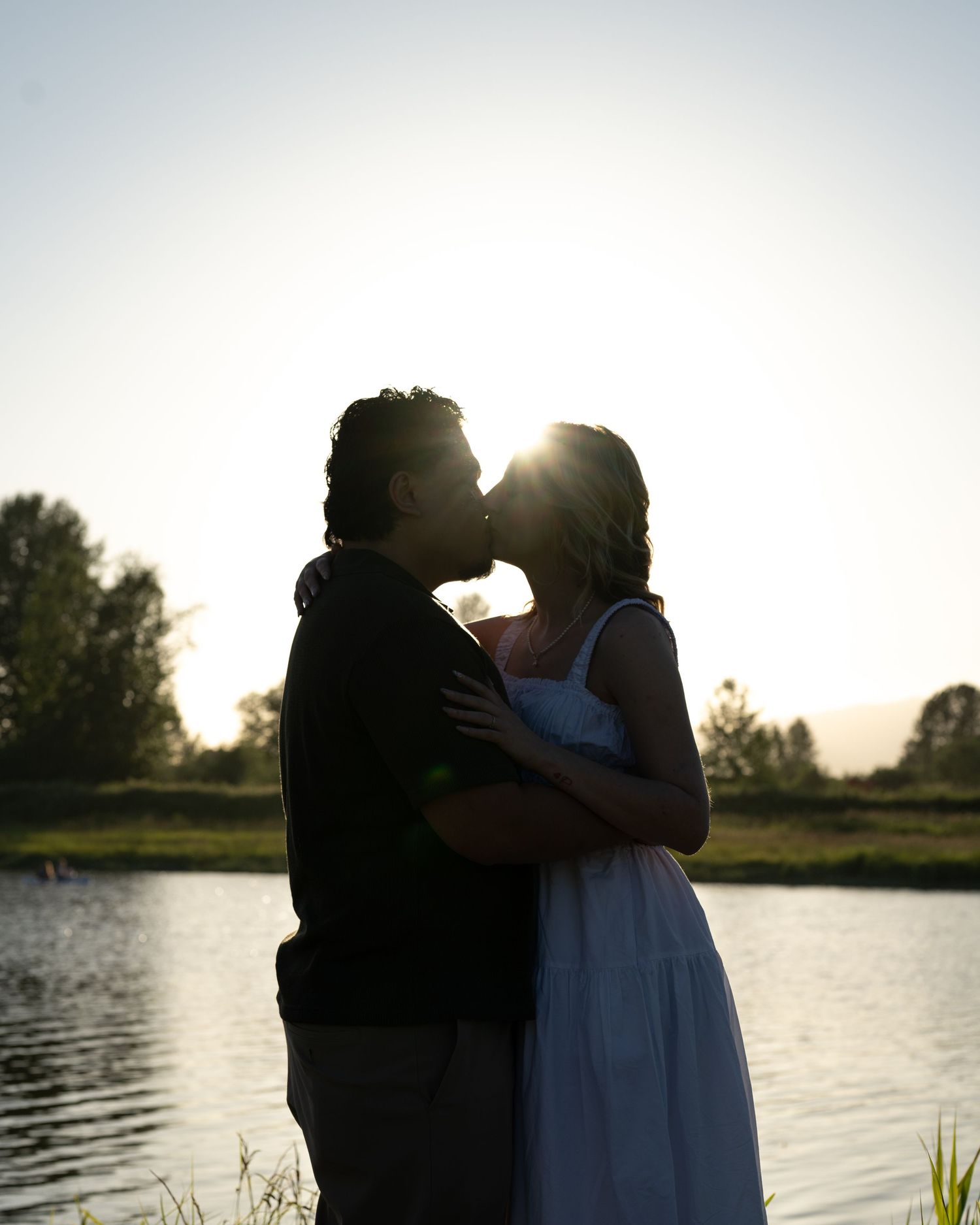 Romantic couple silhouetted against golden sunset light sharing a kiss by a tranquil lake surrounded by trees.