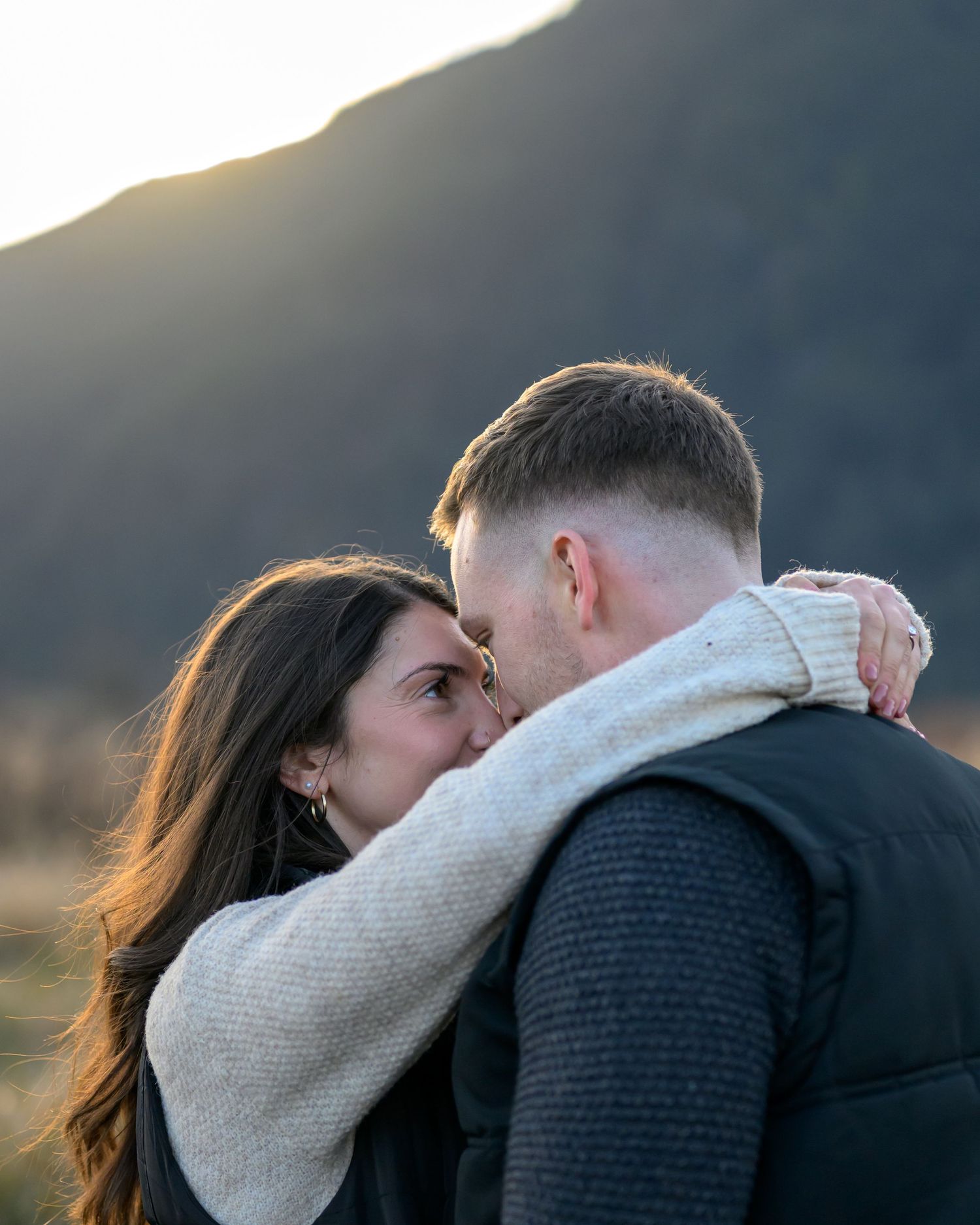 A couple shares a romantic moment outdoors with golden mountain sunset light behind them.