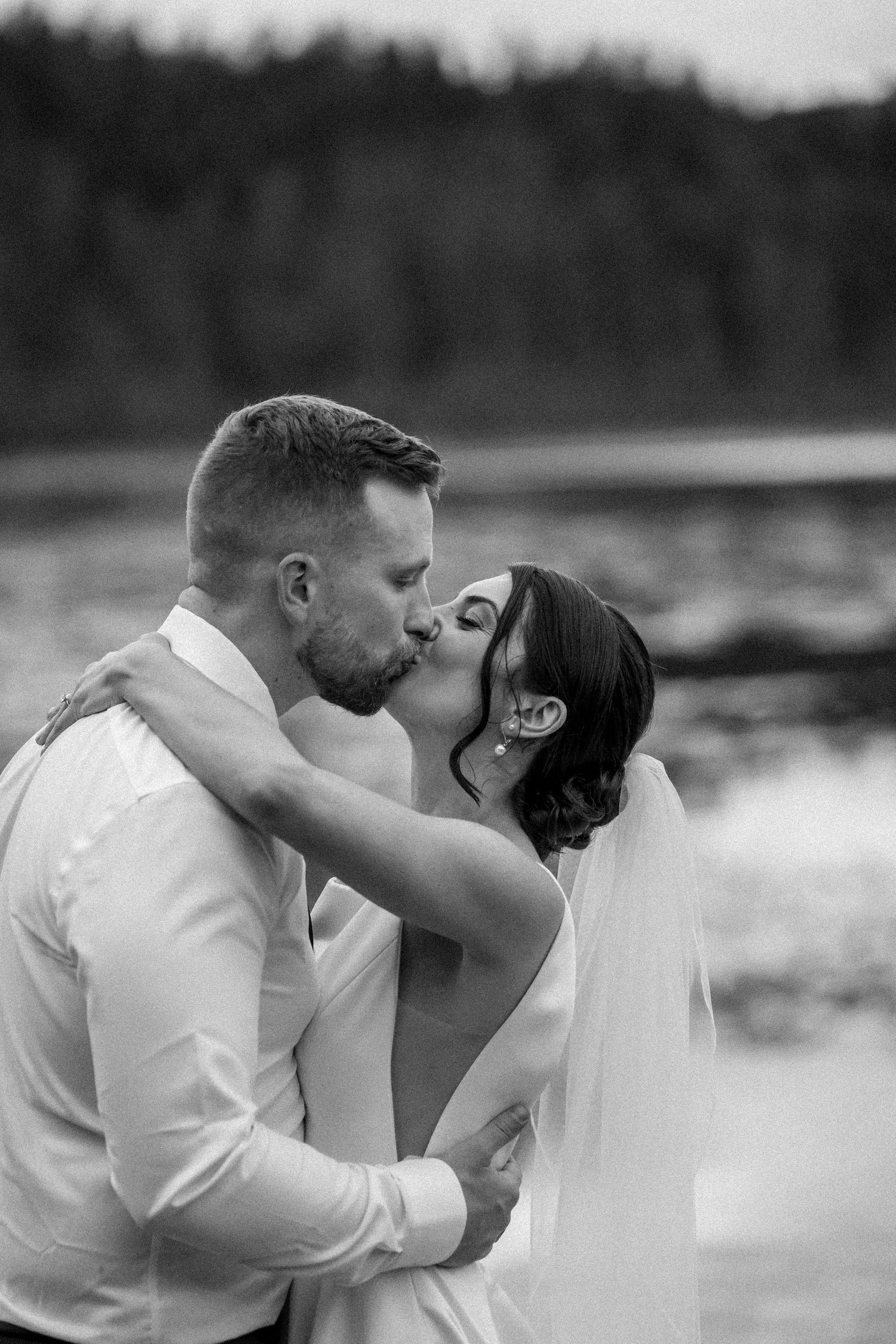 Black and white photo of a bride and groom sharing a romantic kiss by a lake with forest in the background.