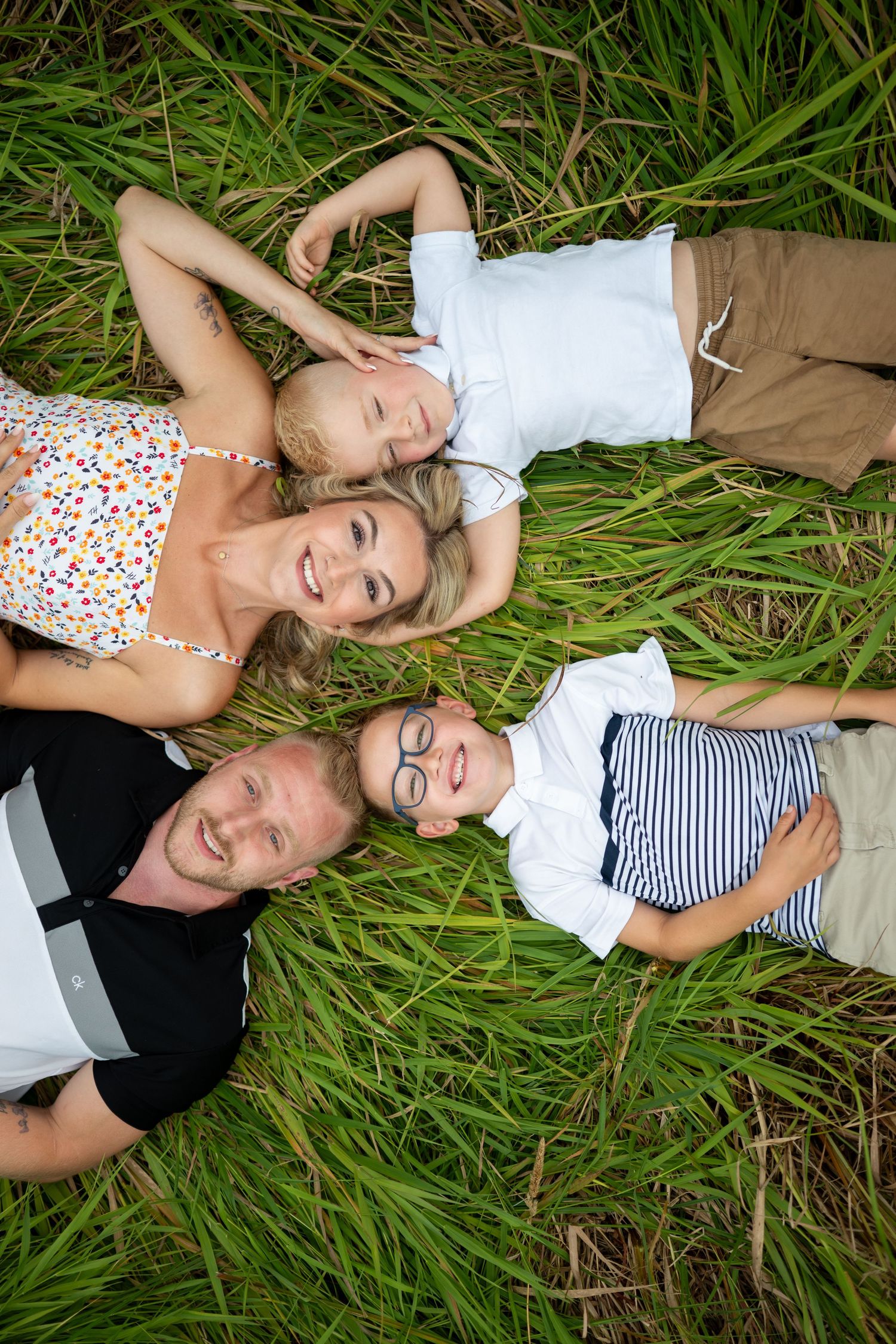 Happy family of four lying in green grass, smiling up at camera in a cheerful outdoor portrait.