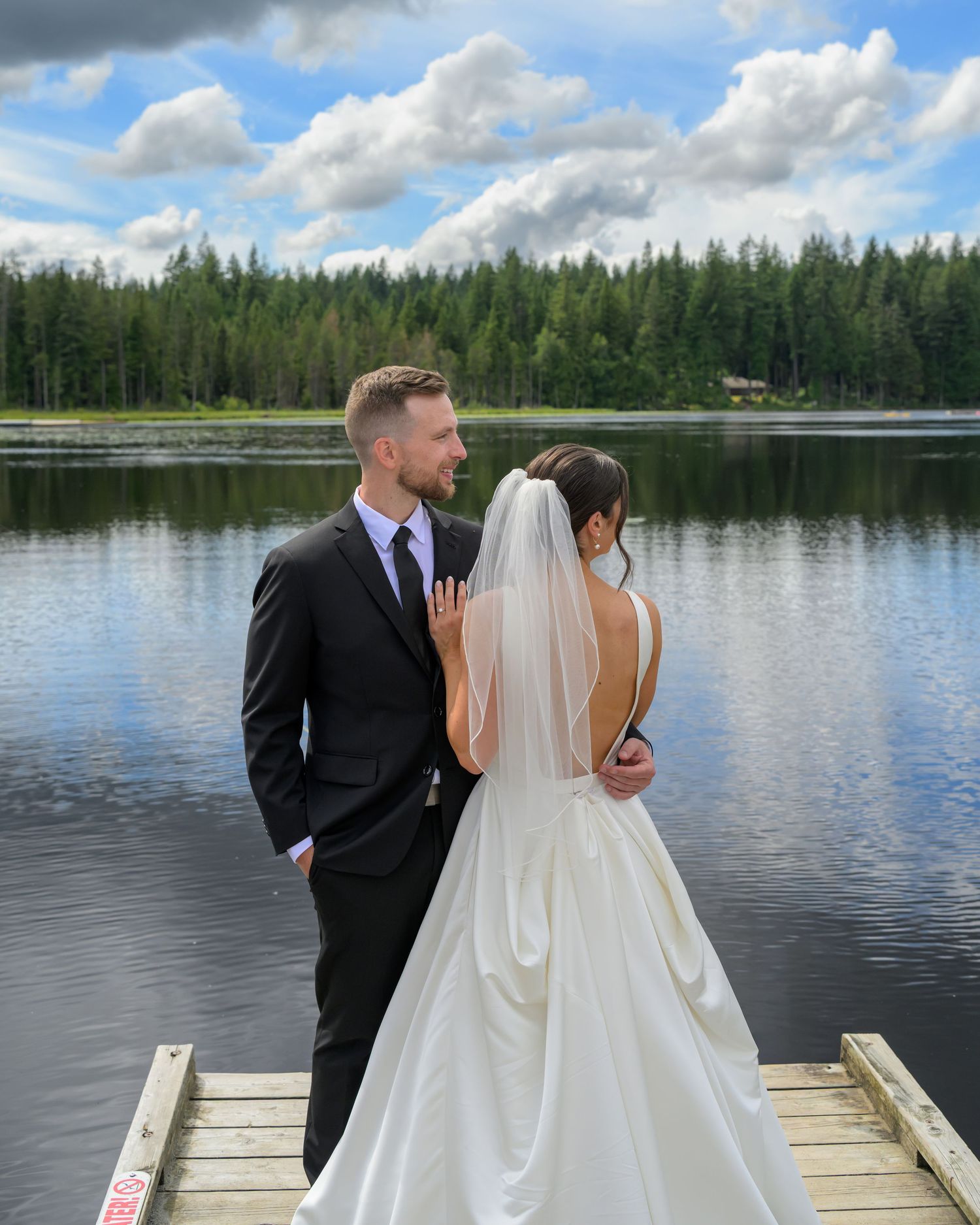 A bride and groom pose on a lakeside dock surrounded by lush forest under a cloudy blue sky.