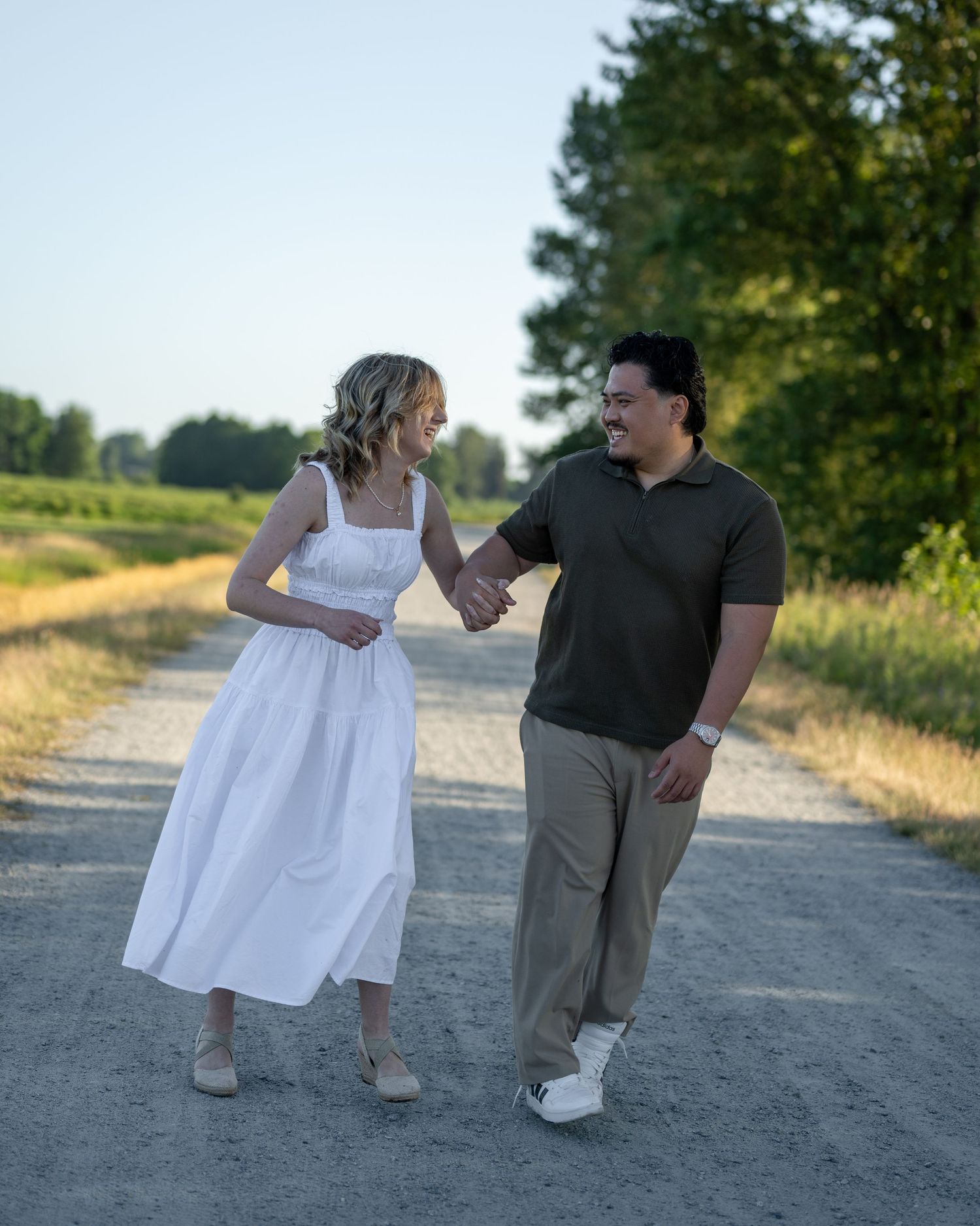 Couple holding hands and smiling while walking together on a sunny gravel path surrounded by green trees and fields.