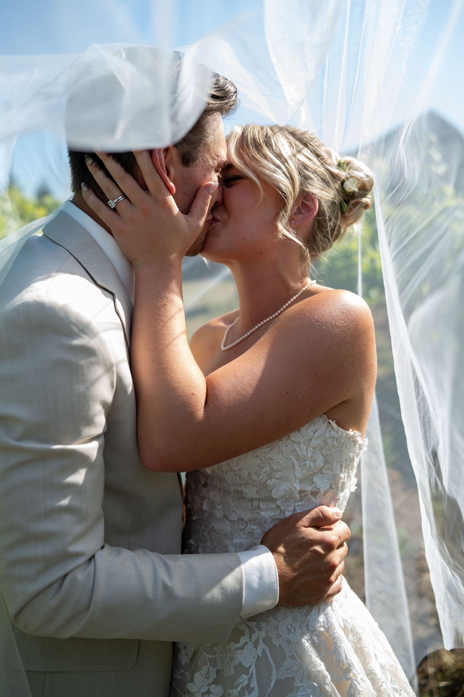 Bride and groom sharing a romantic kiss under a flowing white veil outdoors on their wedding day.