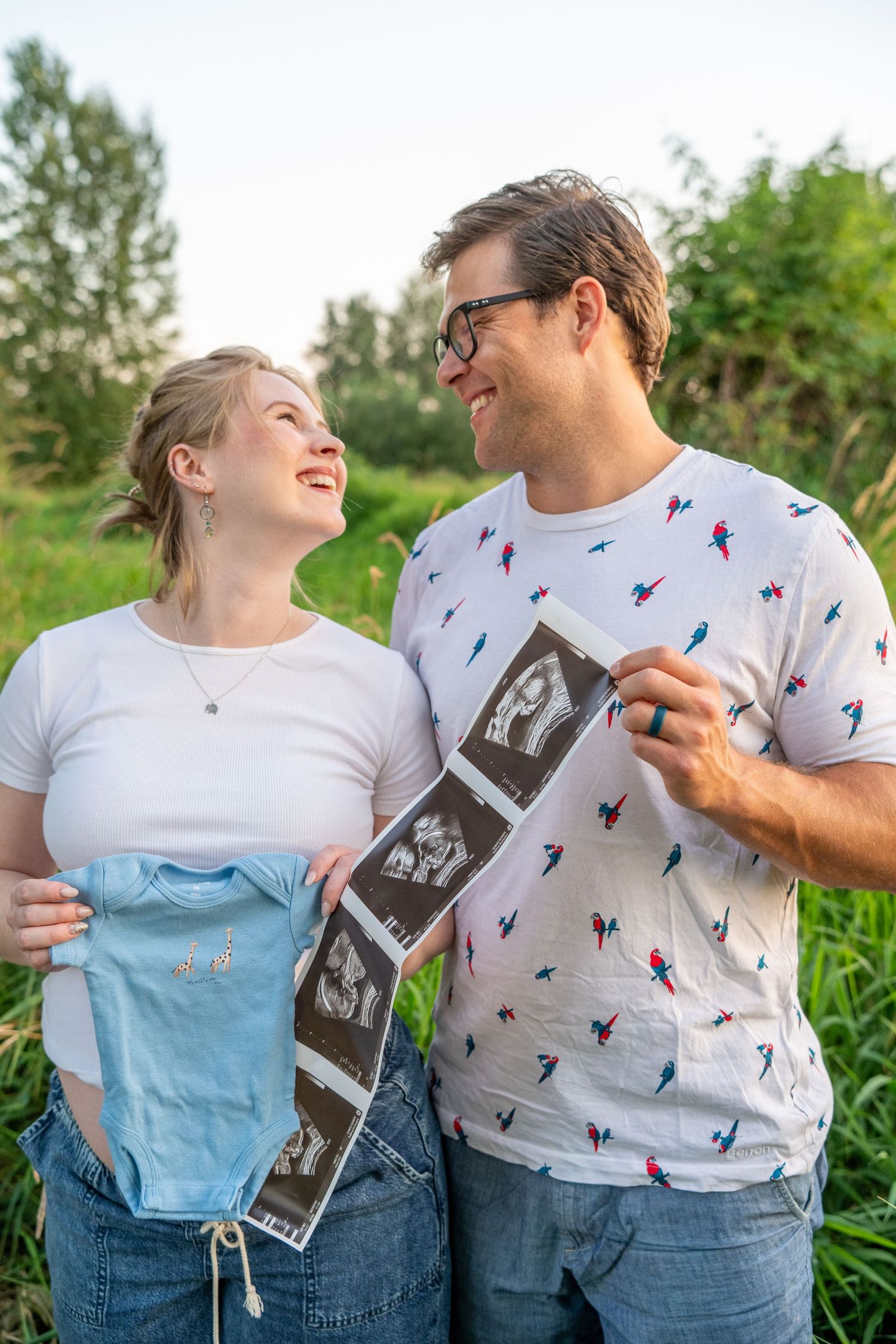 Happy couple smiling at each other outdoors, holding baby onesie and ultrasound scans, announcing pregnancy.