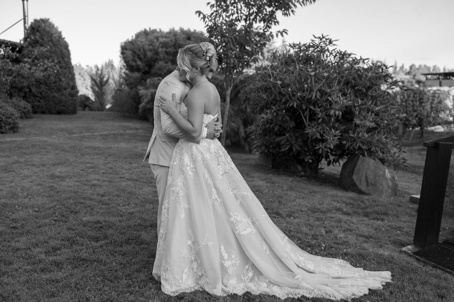 Black and white photo of a bride and groom embracing outdoors on a lawn with lush green trees in the background.