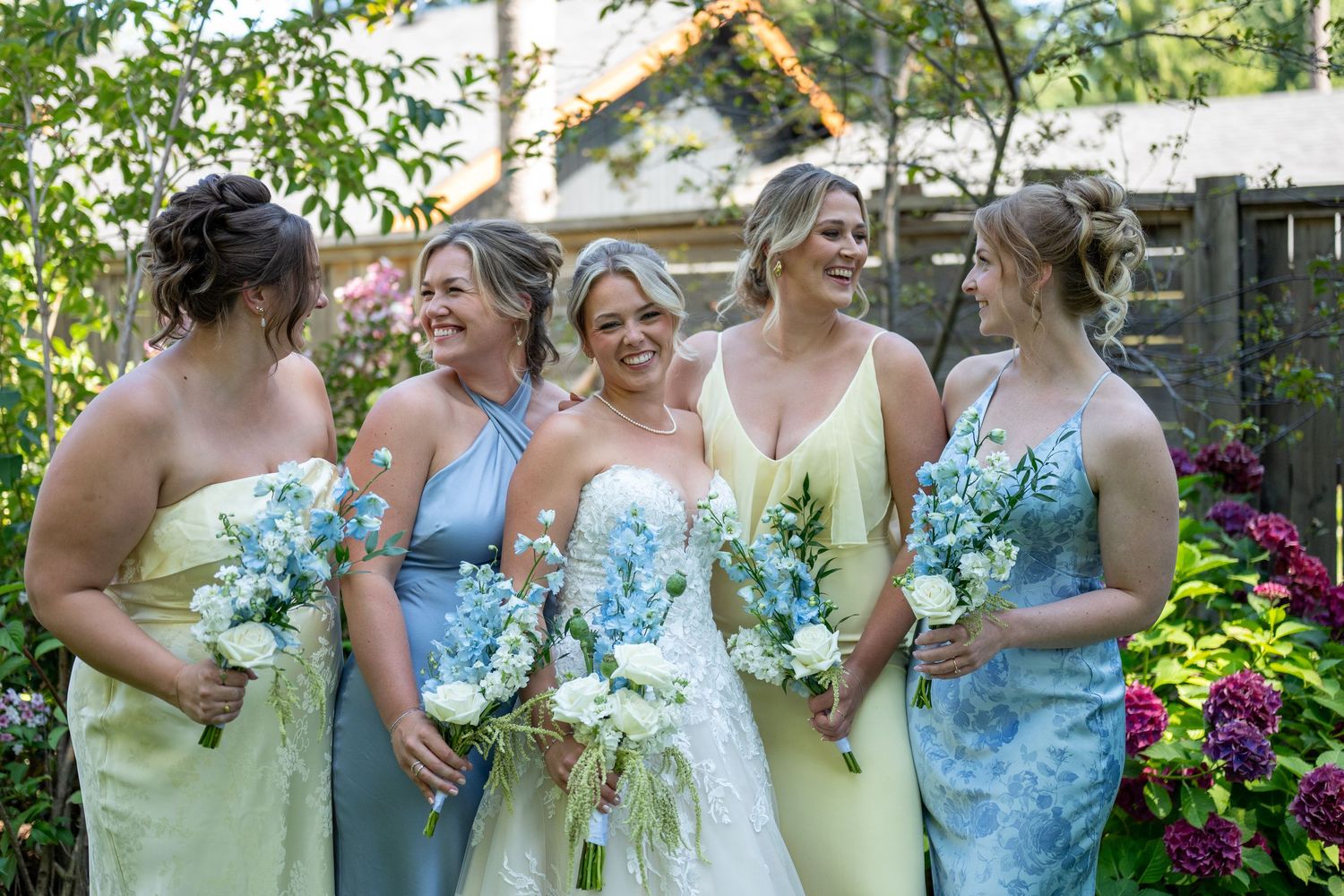 Bride in white gown with four bridesmaids in yellow and blue dresses holding floral bouquets in a garden setting.