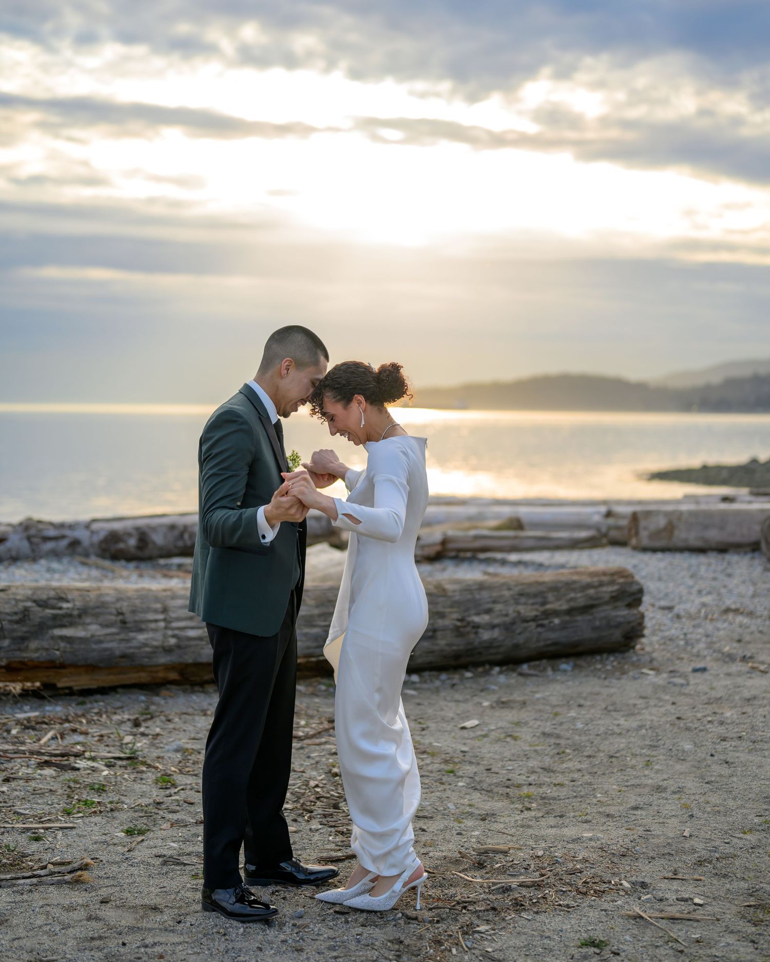Wedding couple dances on driftwood-lined beach during sunset.