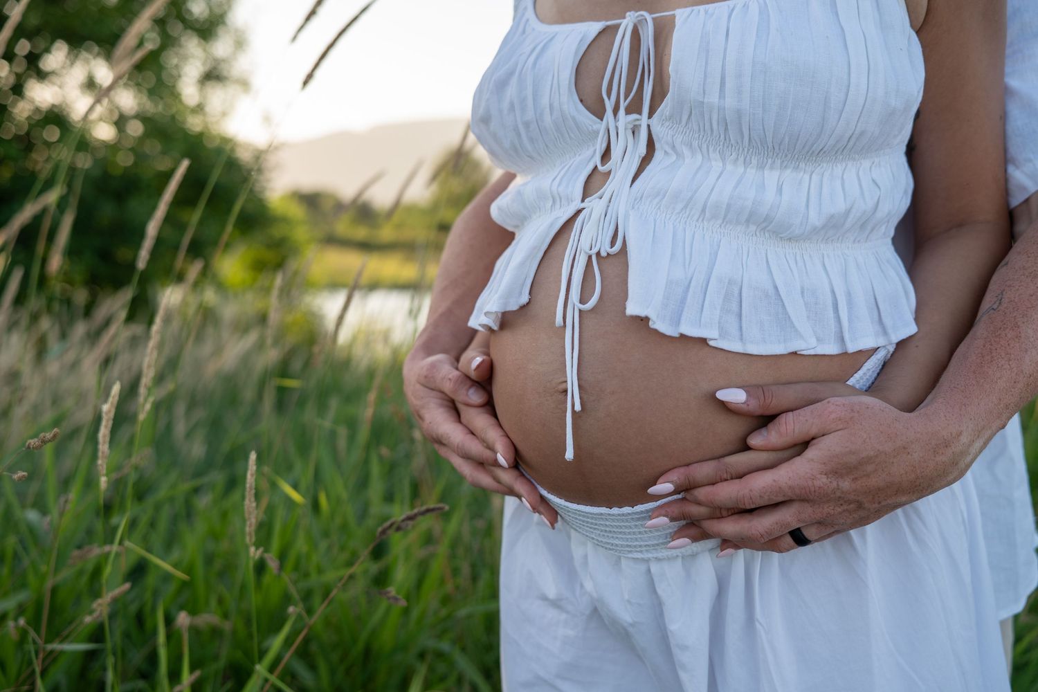 Pregnant woman in white crop top cradling baby bump outdoors near lush green grass and water.