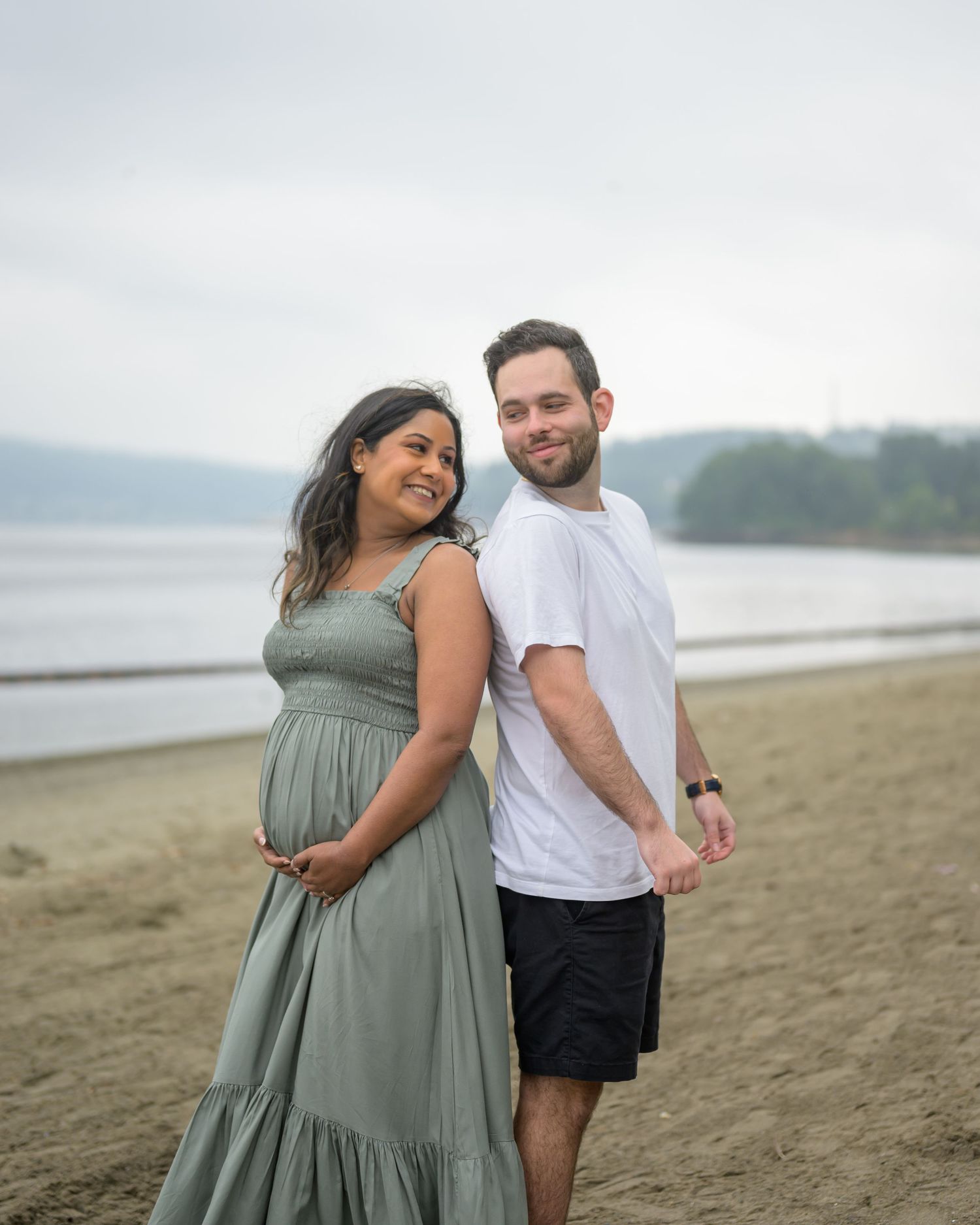 Expecting couple in matching sage outfits smiling on sandy beach during maternity photoshoot.