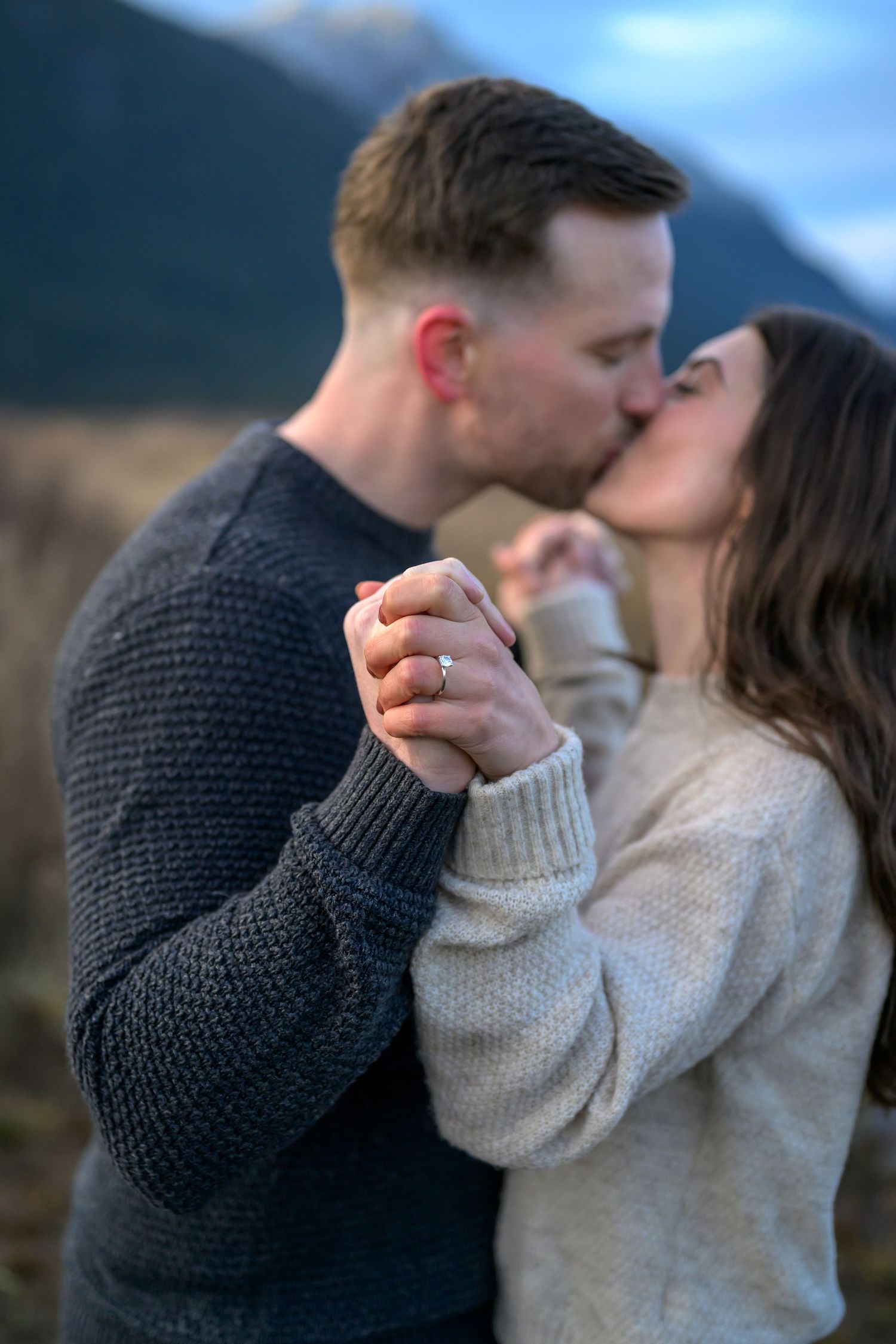 Couple sharing a romantic kiss outdoors, holding hands with engagement ring visible, mountains in the background.