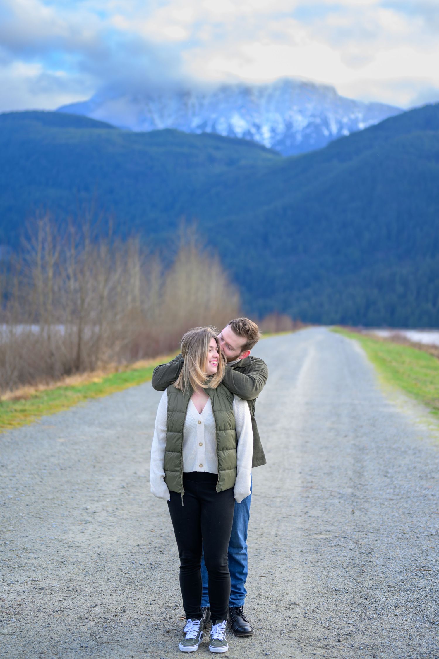 Couple playfully posing on gravel path with snow-capped mountains in background.