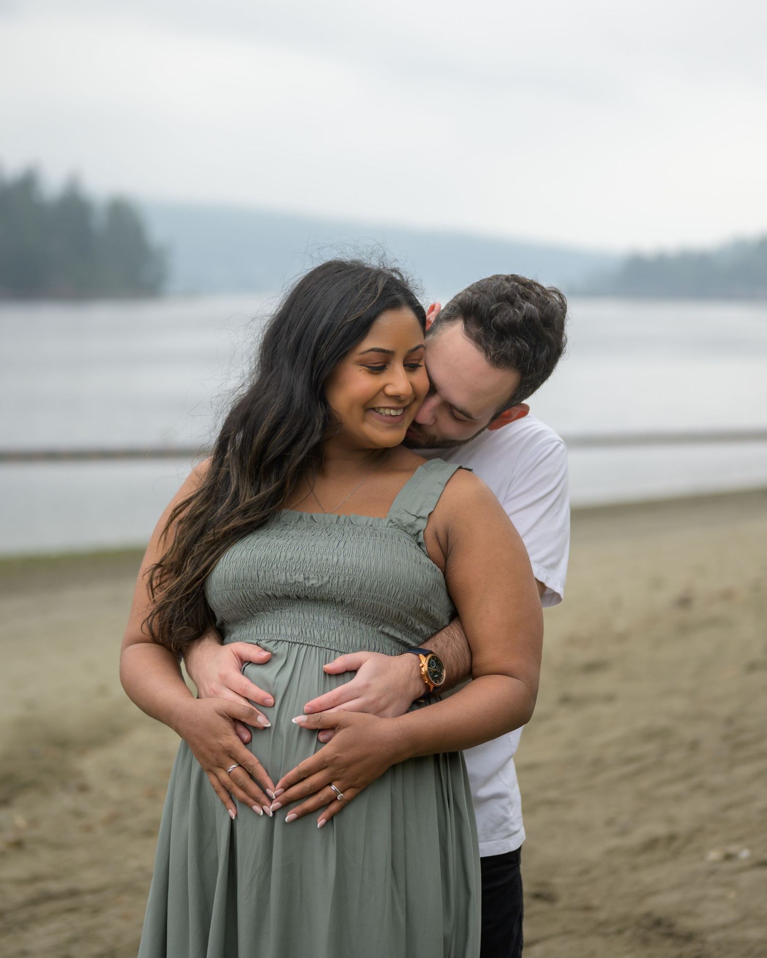 Pregnant woman in sage green dress embraced by partner on beach, both cradling baby bump during maternity photoshoot.