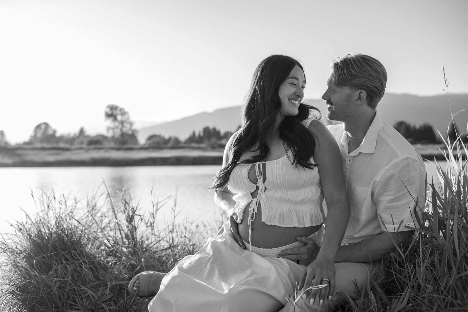 Black and white photo of a smiling couple sitting together by a lake surrounded by tall grass and mountains.
