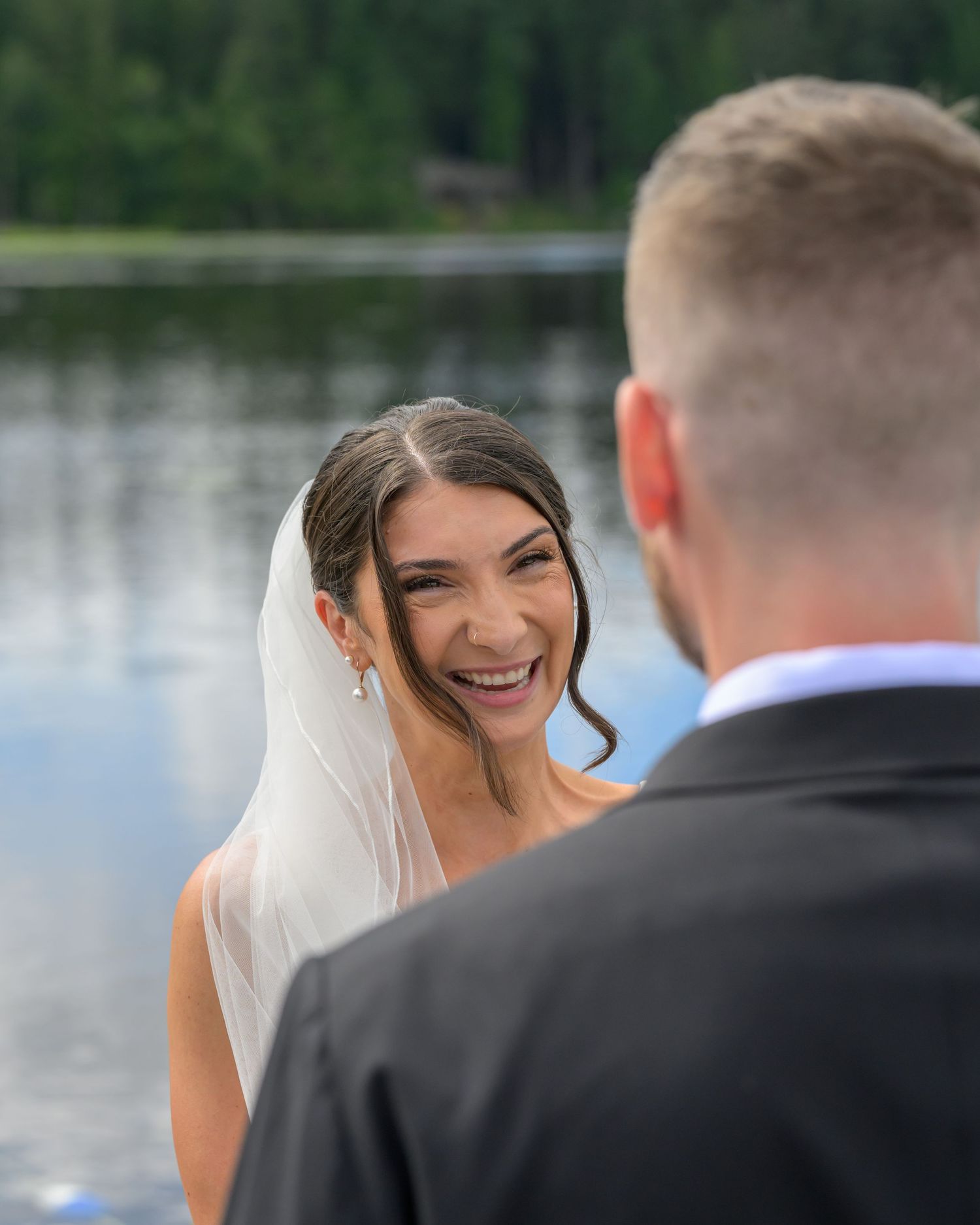 Bride in veil laughing joyfully at groom during outdoor lakeside wedding ceremony with forest backdrop.