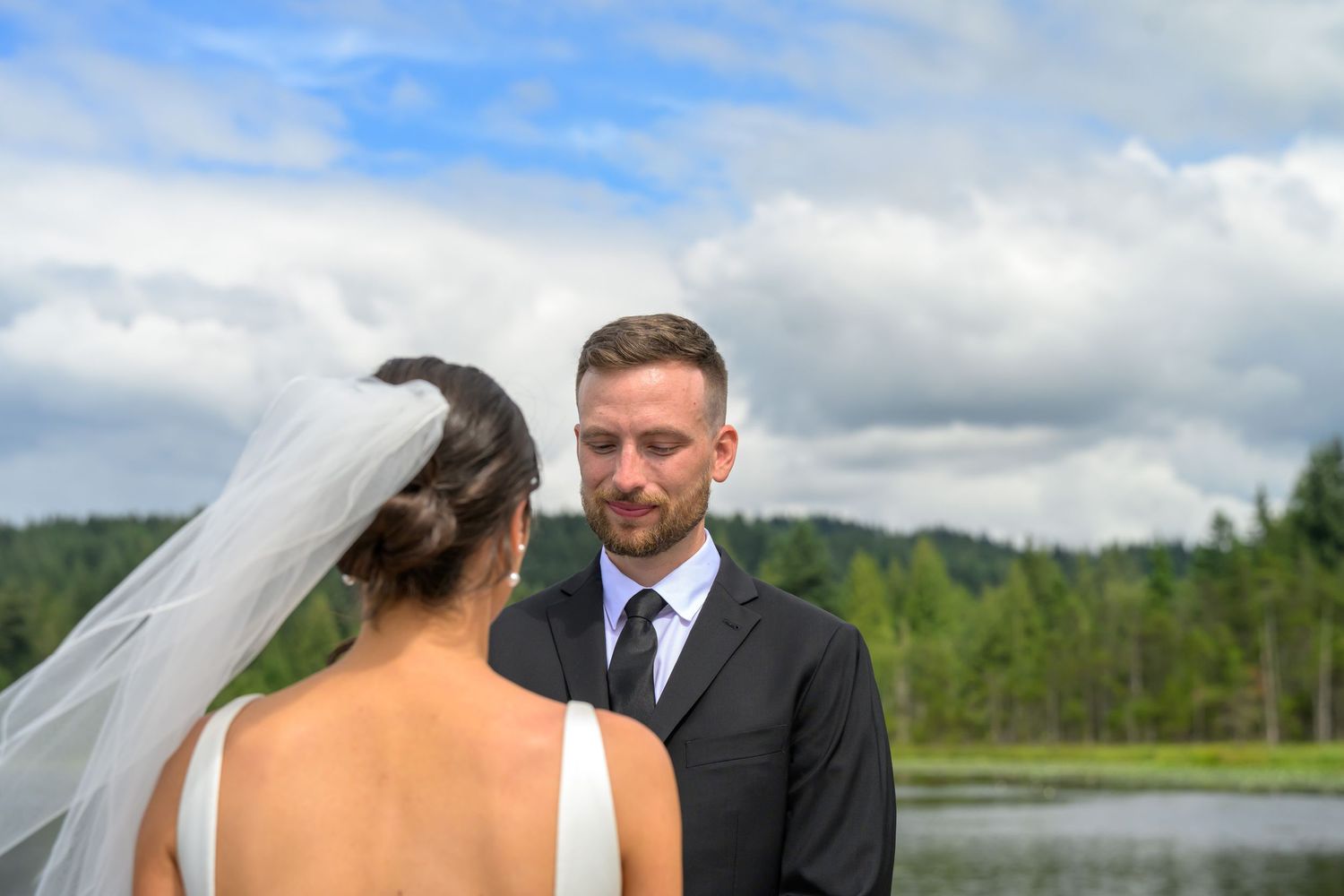 Bride and groom exchange vows outdoors near a lake with lush green trees and cloudy blue sky backdrop.