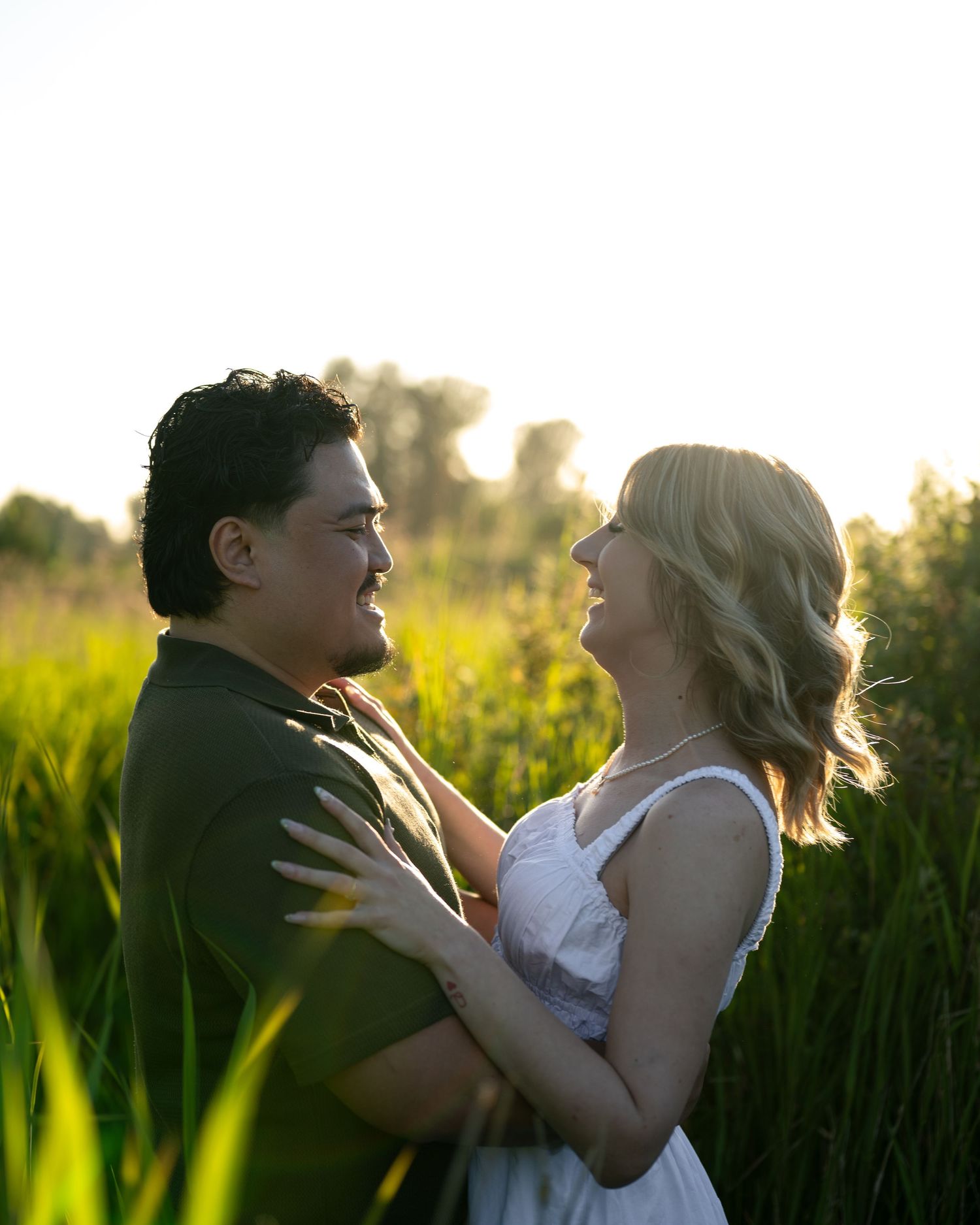 A couple embraces in a sunlit field, gazing at each other surrounded by lush green grass at golden hour.