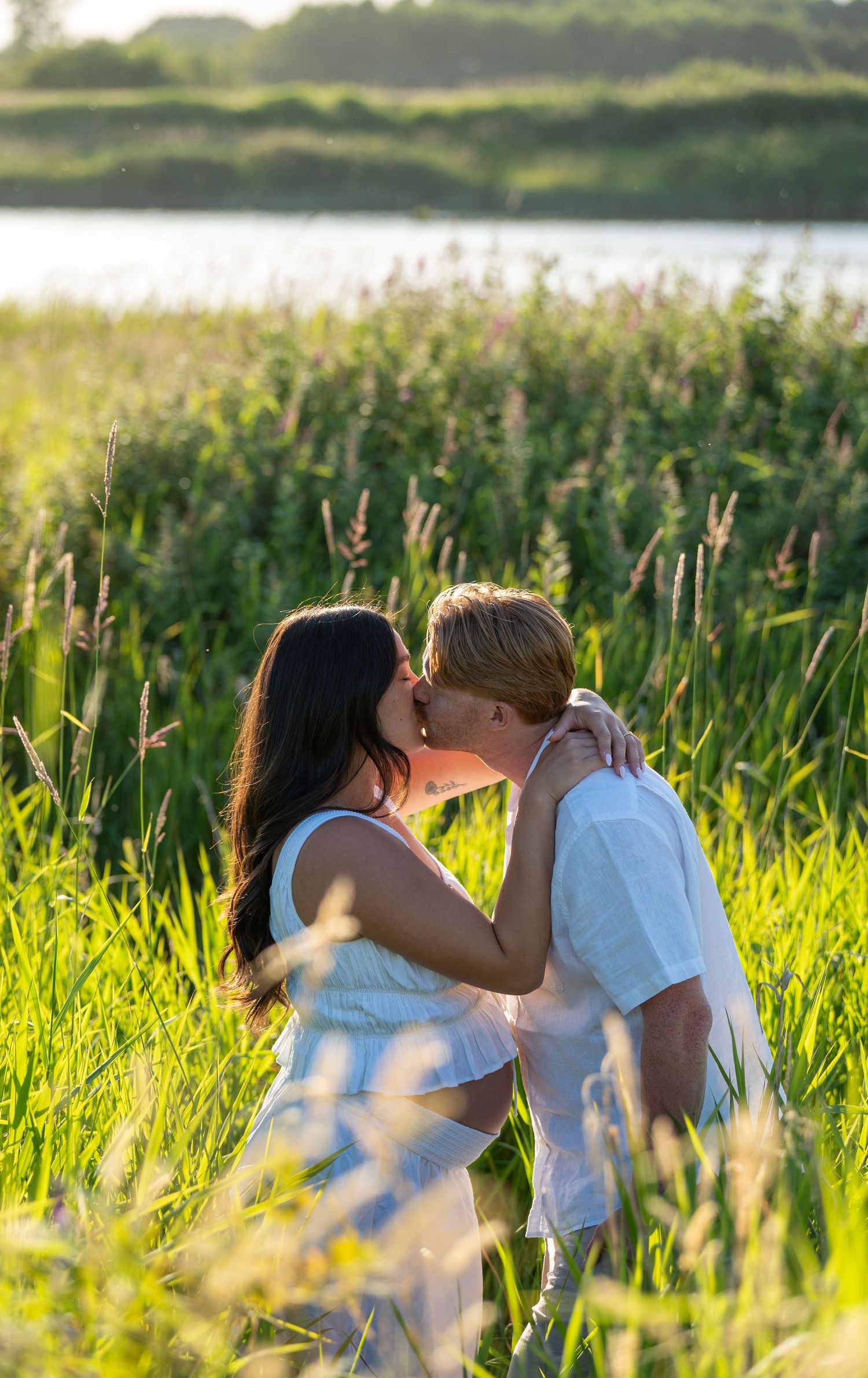 Couple sharing a romantic kiss in a lush green field with a serene lake visible in the background.