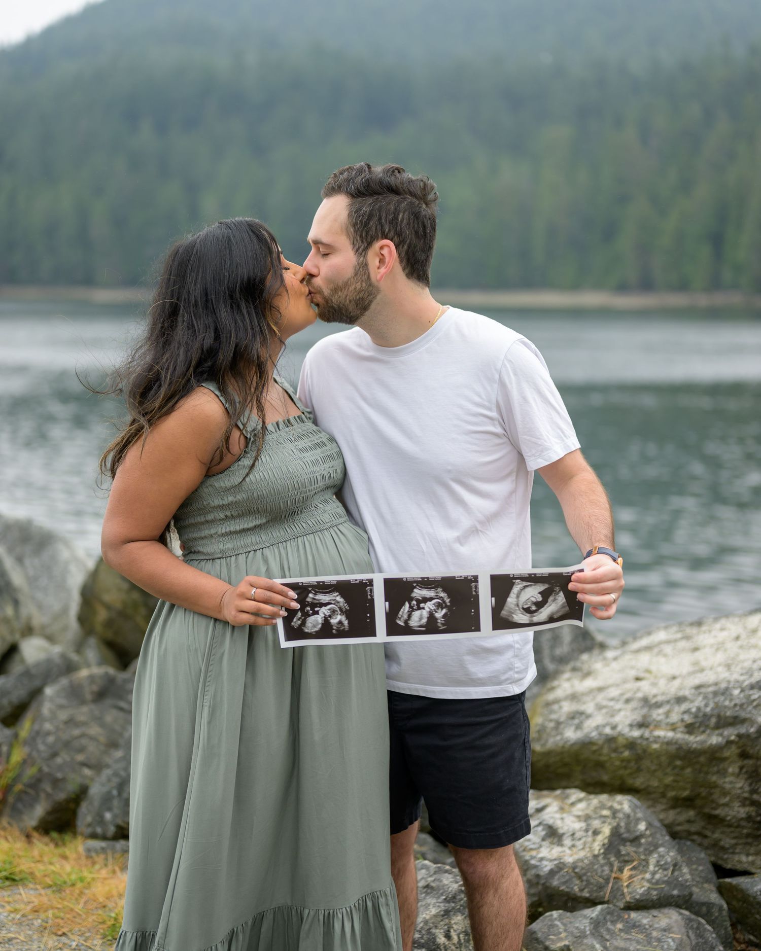Couple kissing outdoors by a lake holding ultrasound photos, announcing pregnancy, with mountains in the background.