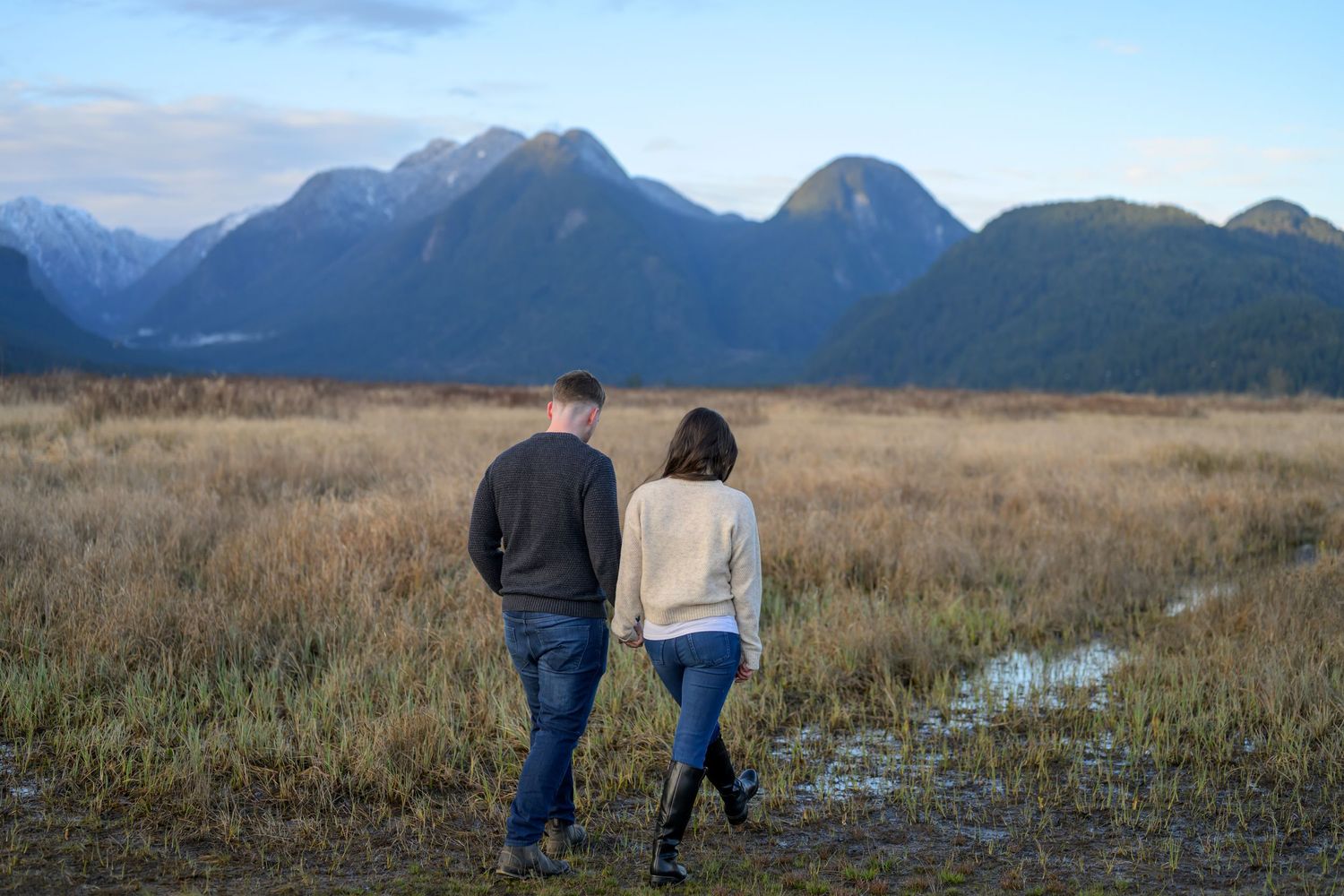 Couple walking hand in hand through a golden marsh field with stunning mountain backdrop at dusk.