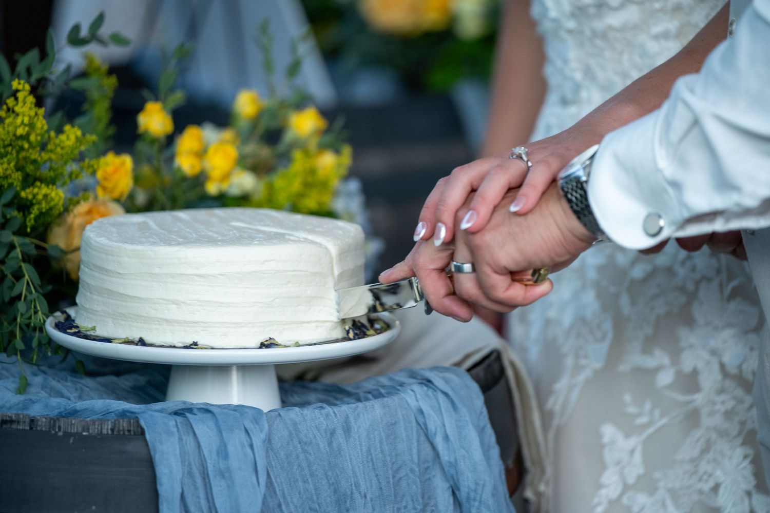 Bride and groom cutting a white wedding cake on a pedestal stand with yellow flowers in the background.