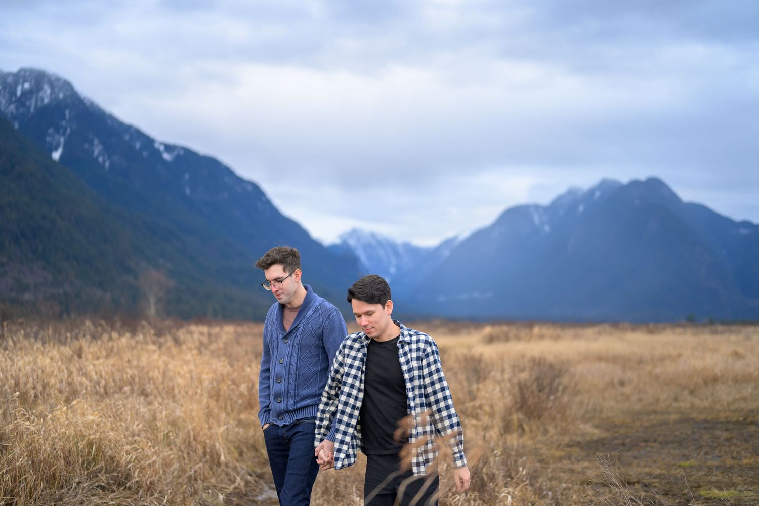 Couple walking through golden field with mountain range backdrop.