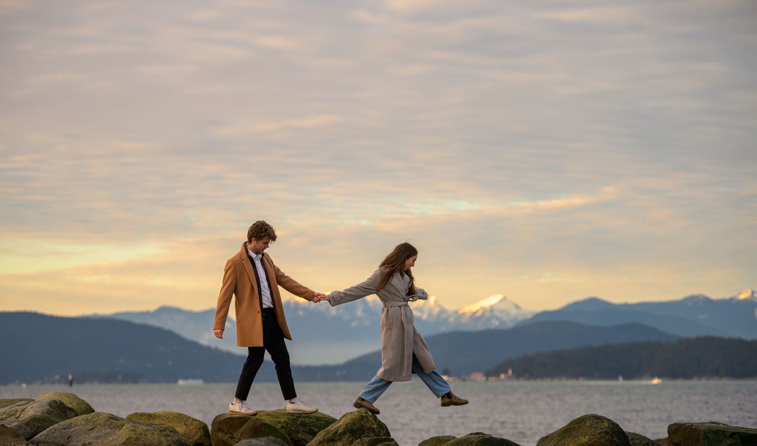 Silhouettes of people dancing on a rocky beach at sunset with mountains in the background.