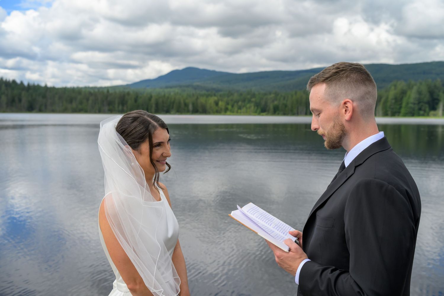 Bride and groom exchange vows lakeside with mountain backdrop on their wedding day.
