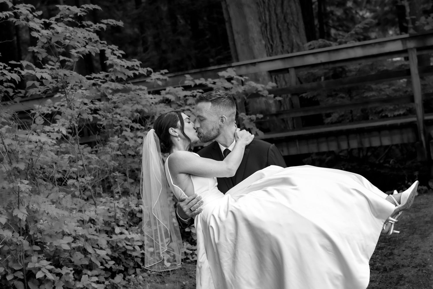 Black and white photo of groom lifting bride for a romantic kiss in a lush garden setting.