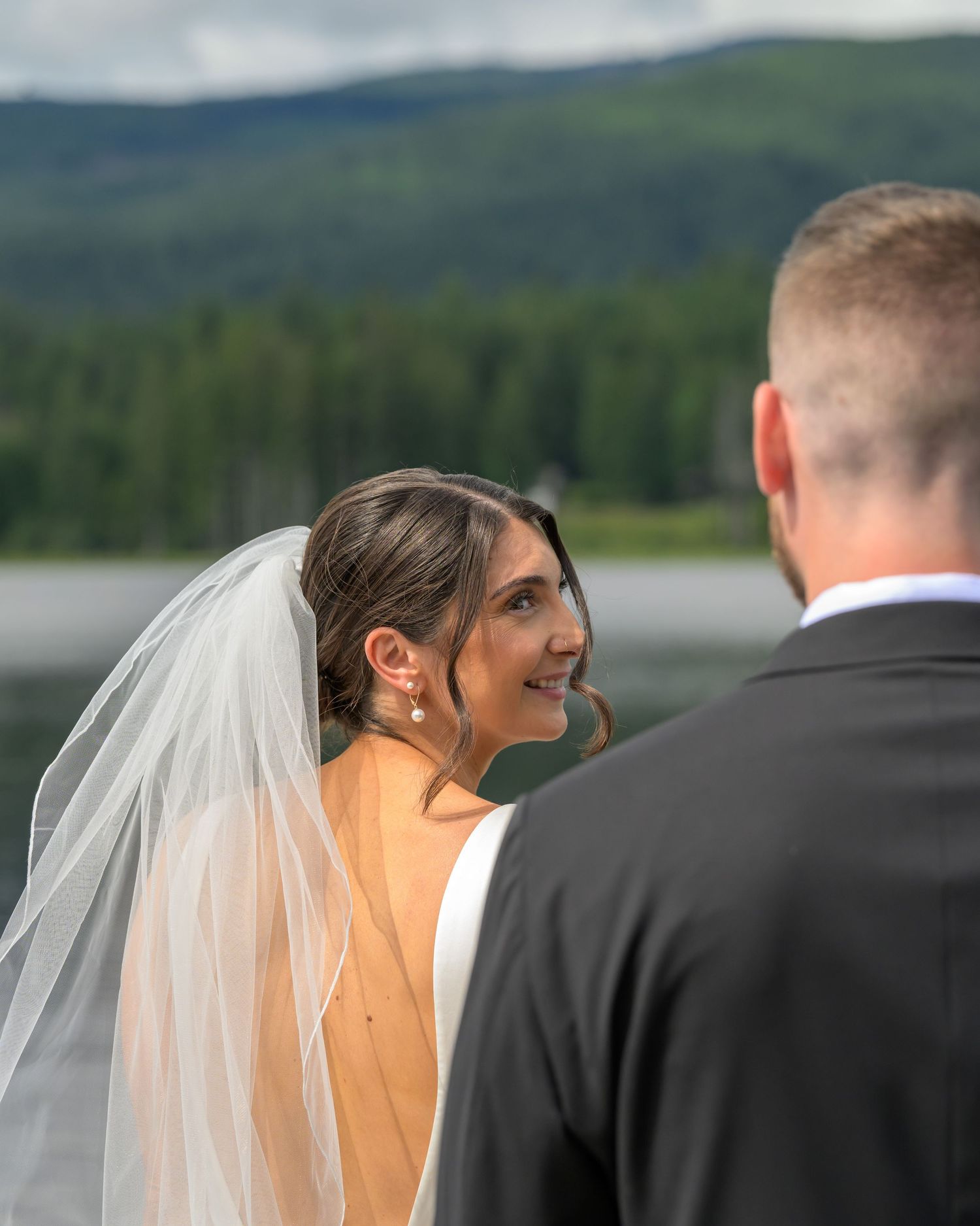 Bride in white veil smiles at groom during outdoor wedding ceremony with mountain lake backdrop.